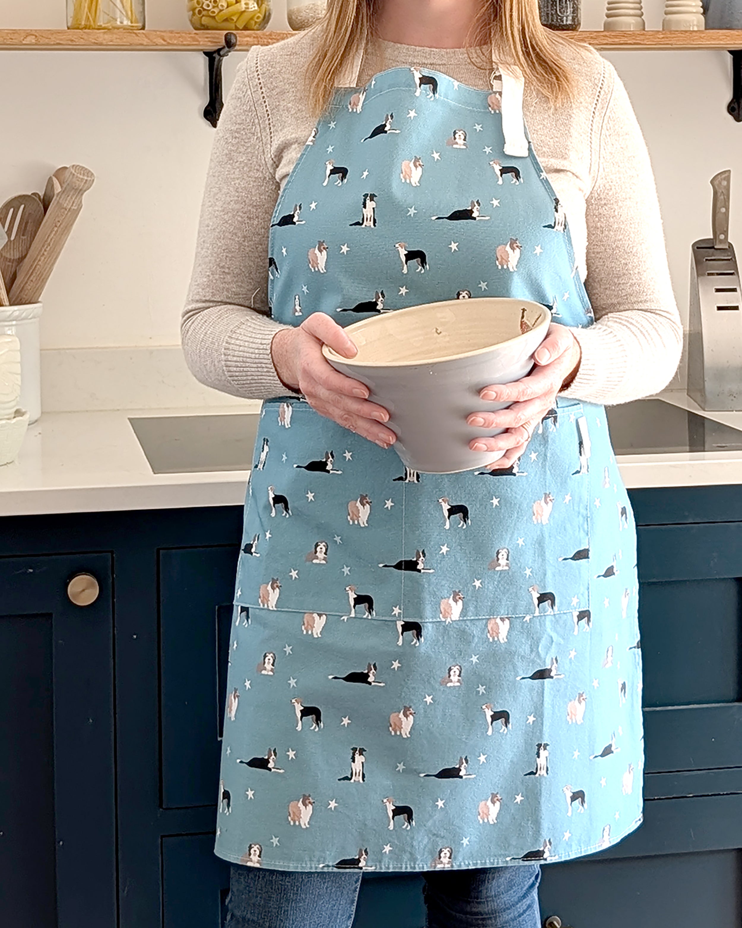 Person wearing a blue apron with collie dog pattern holding a bowl in a kitchen.