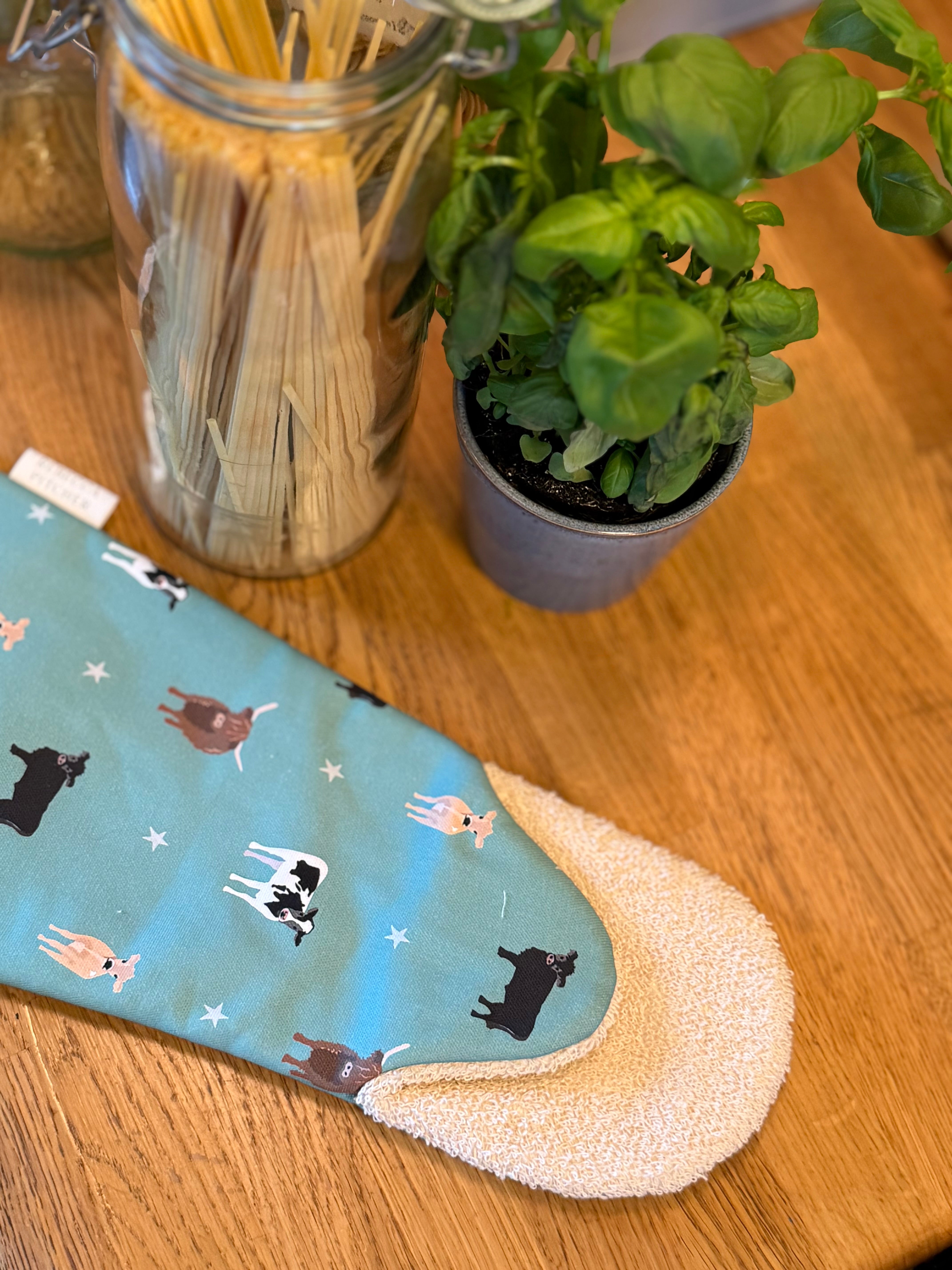 Green cow design oven gauntlet on a wooden worktop with herbs and pasta