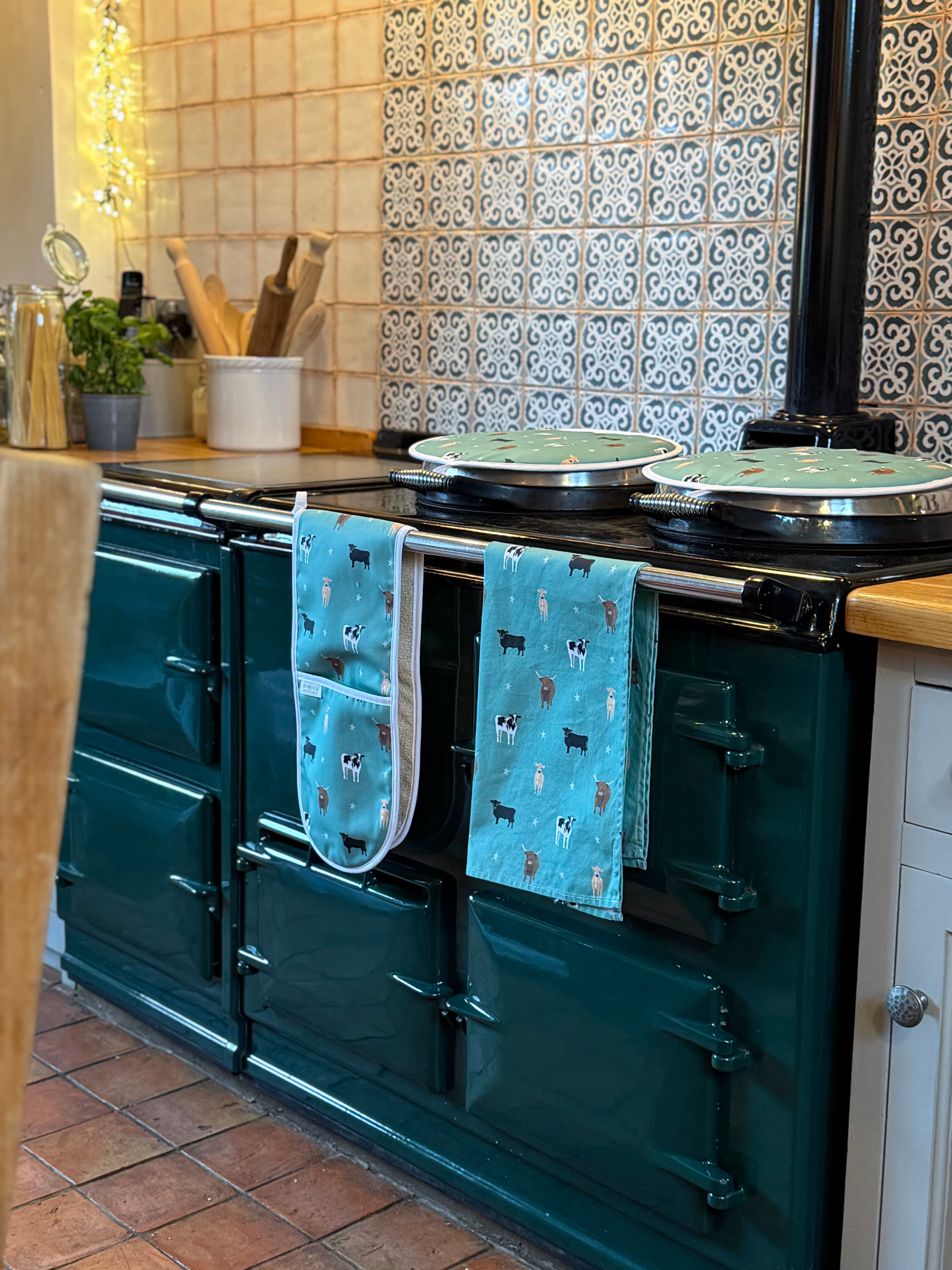 Teal Aga range cooker with green cow print towel, oven gloves and hob covers hanging on the handle in a country kitchen setting.