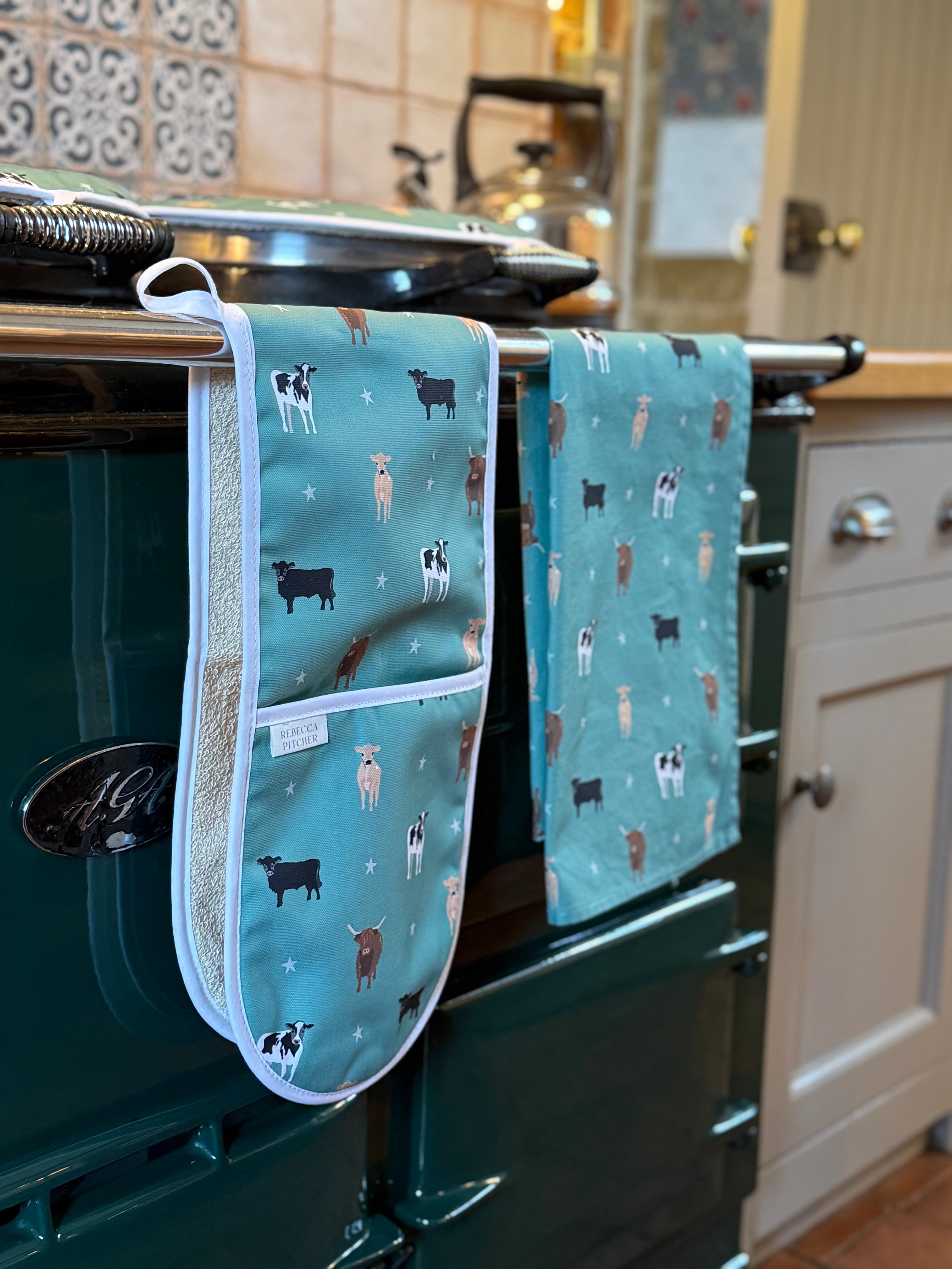 Green cow design oven mitts and tea towels hanging on a kitchen stove.