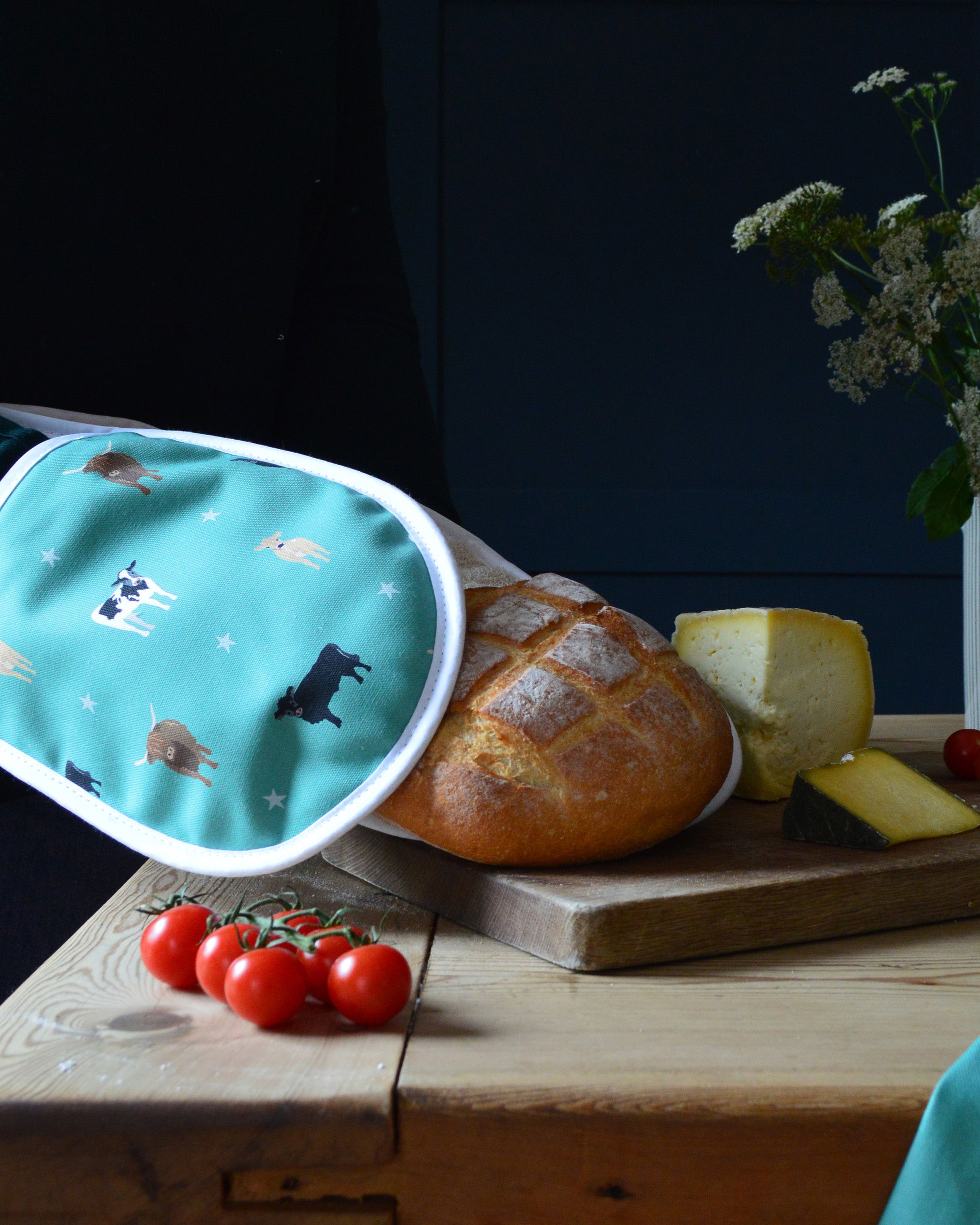 Green oven gloves with cow pattern on a wooden table with bread and tomatoes.