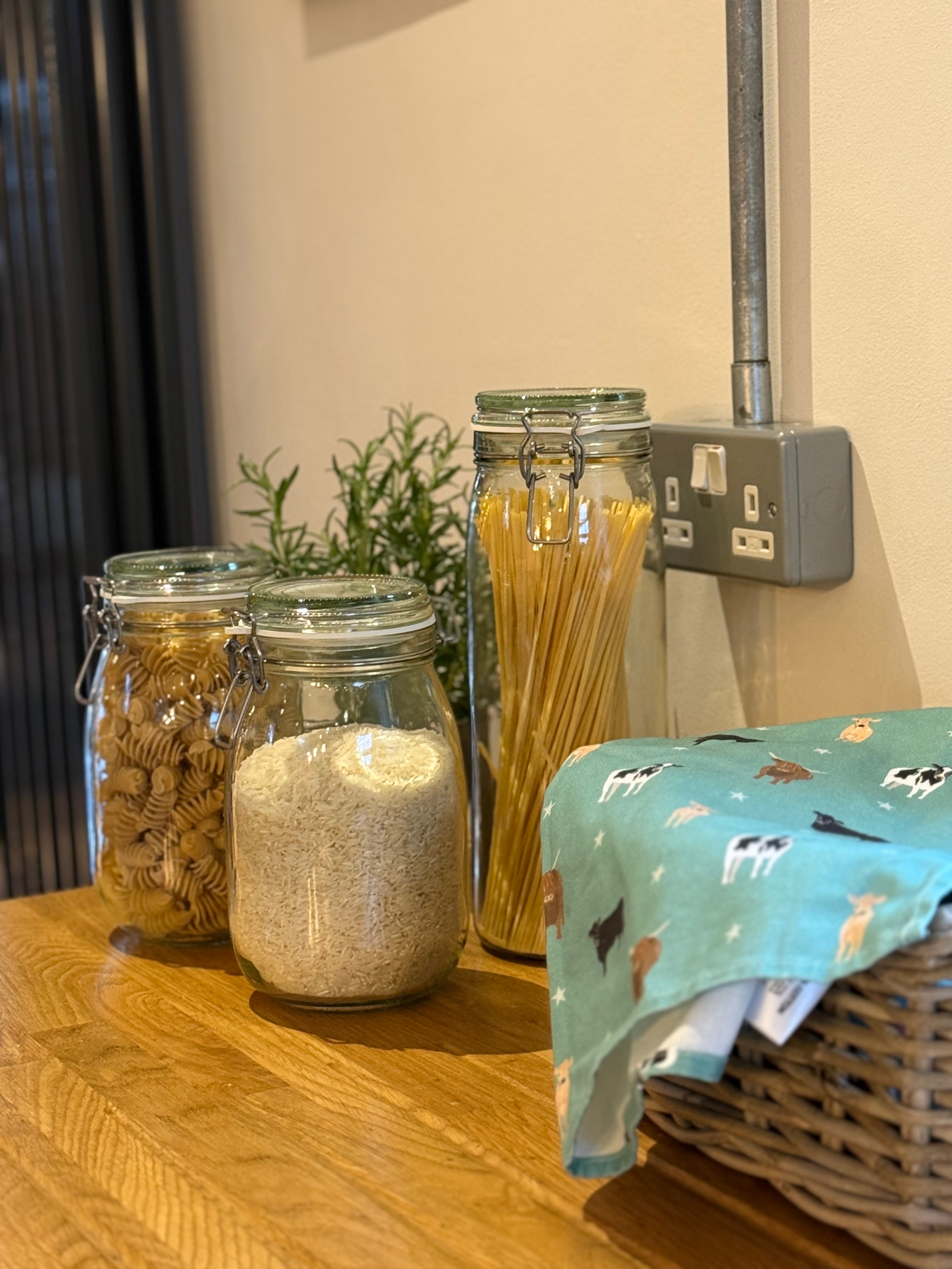 Green cow design tea towel in a basket with jars of pasta and flour on a wooden surface in the background.