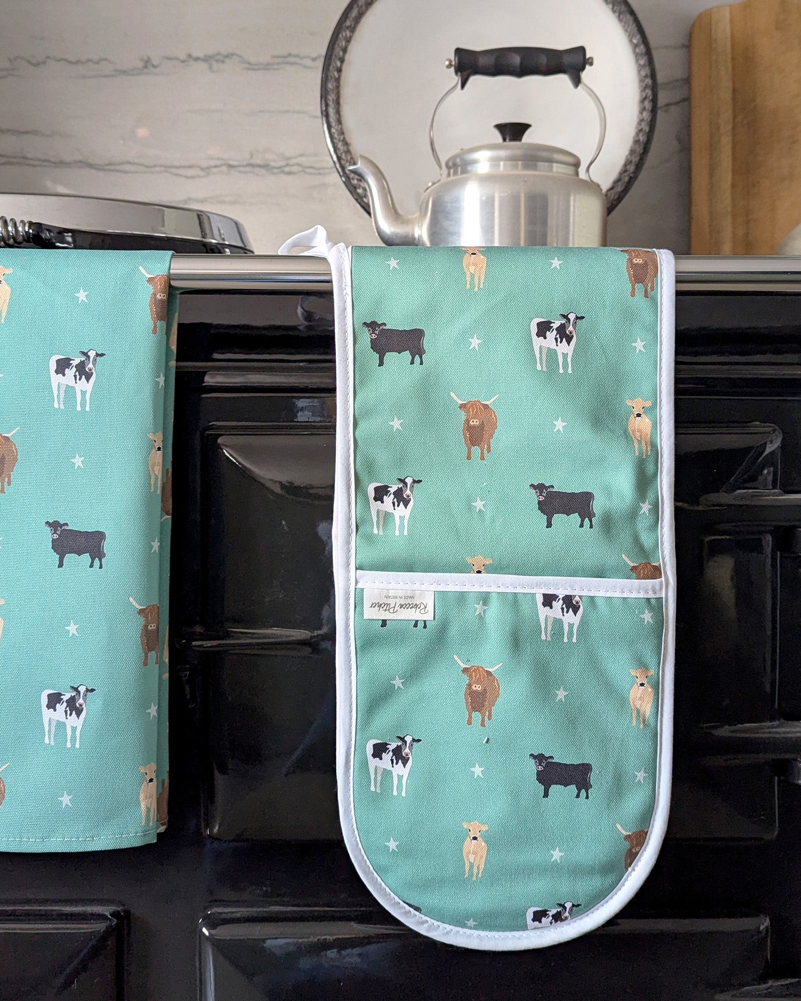 Green oven mitt and tea towel with cow pattern on a kitchen stove.