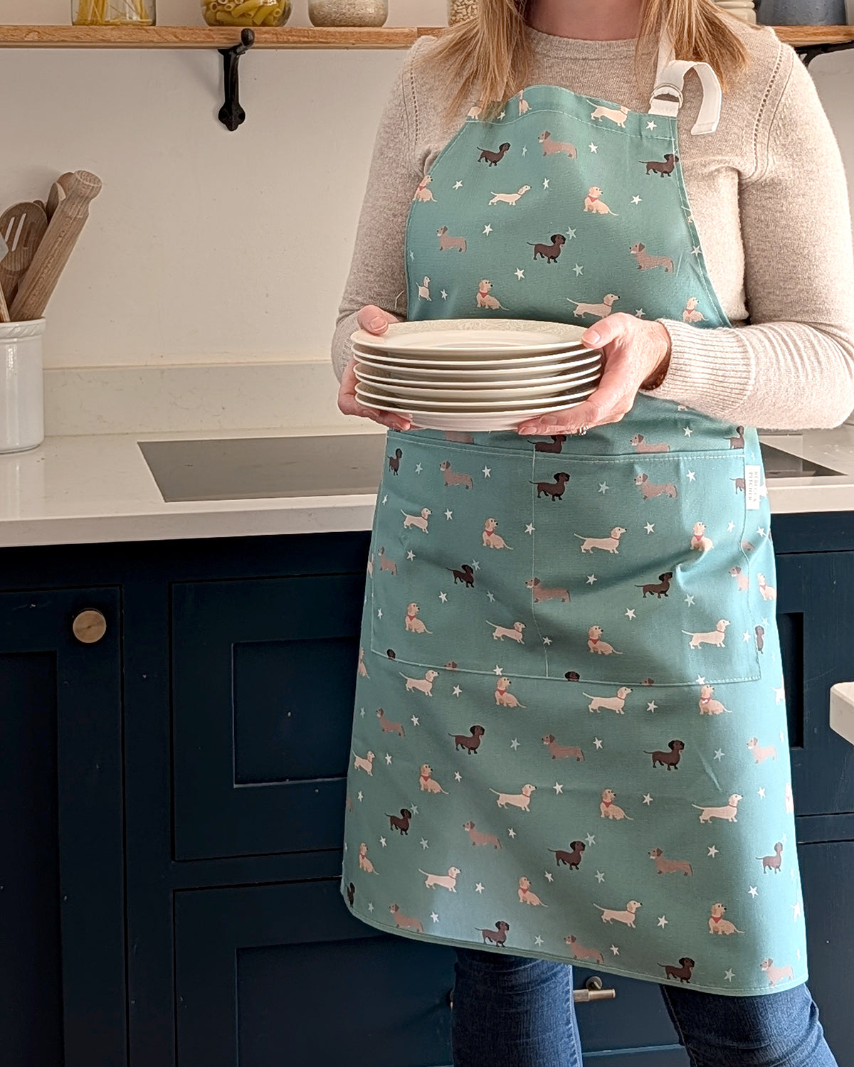Person wearing a green apron with sausage dog pattern holding plates in a kitchen.
