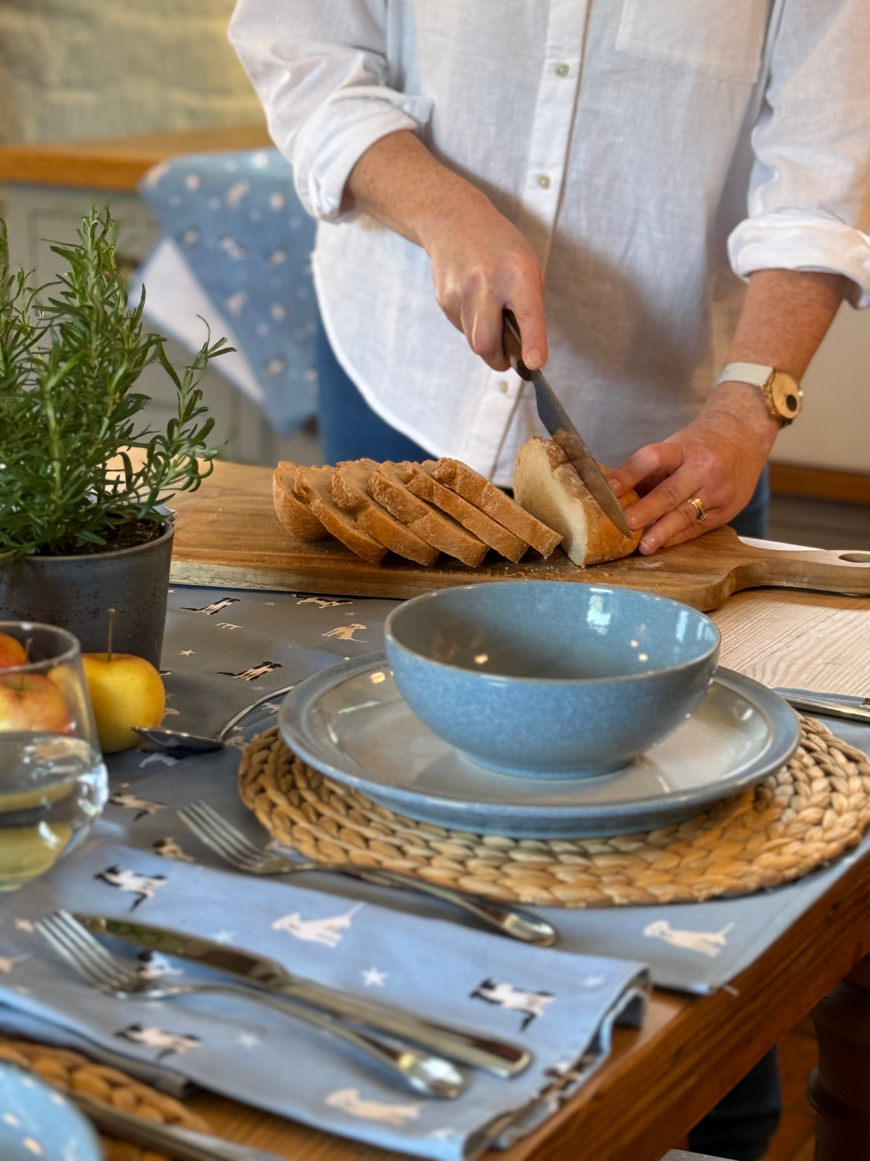 Person slicing bread on a wooden cutting board with a table setting in the foreground