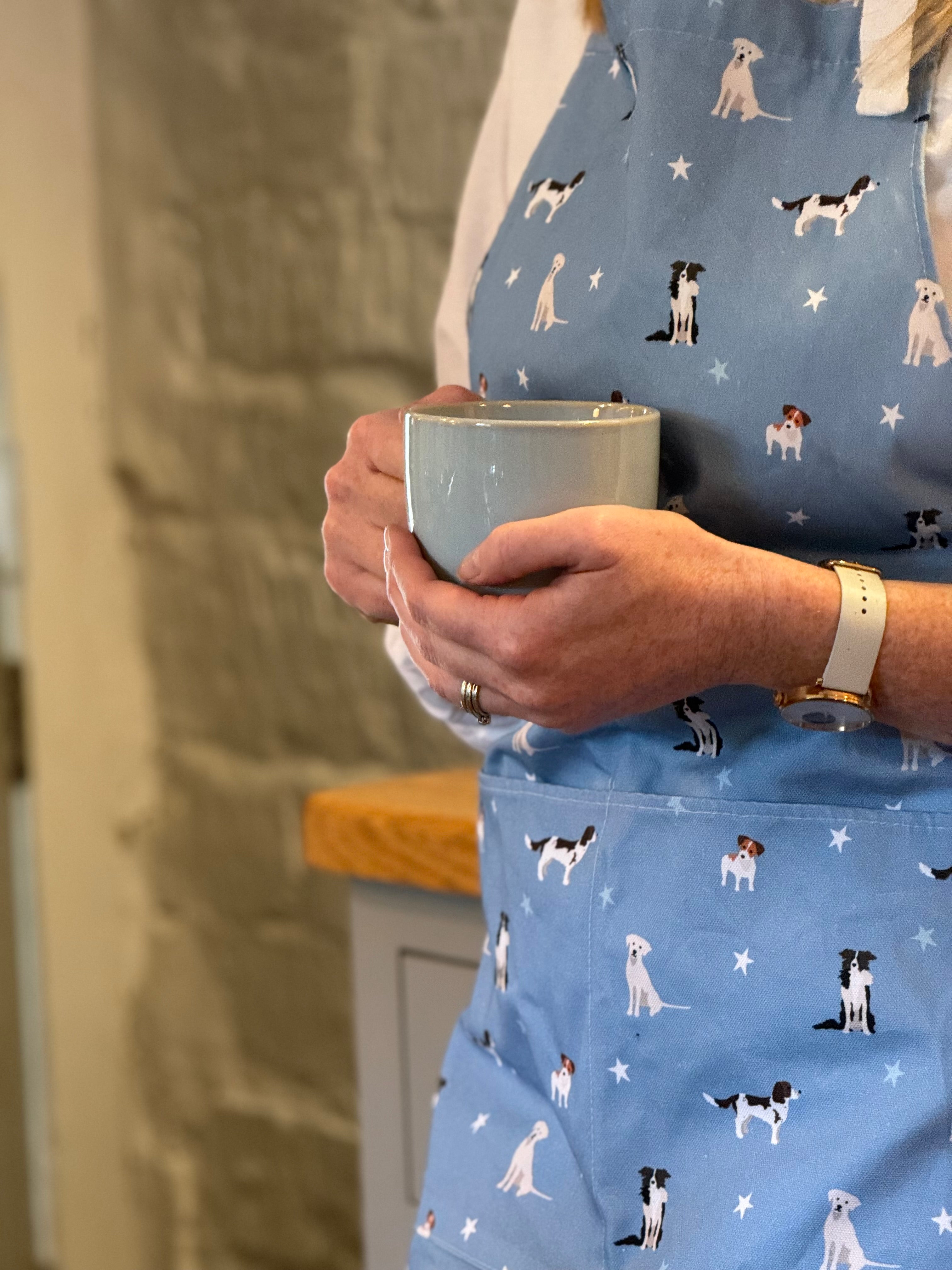 Person wearing a blue apron with dog patterns, holding a mug.