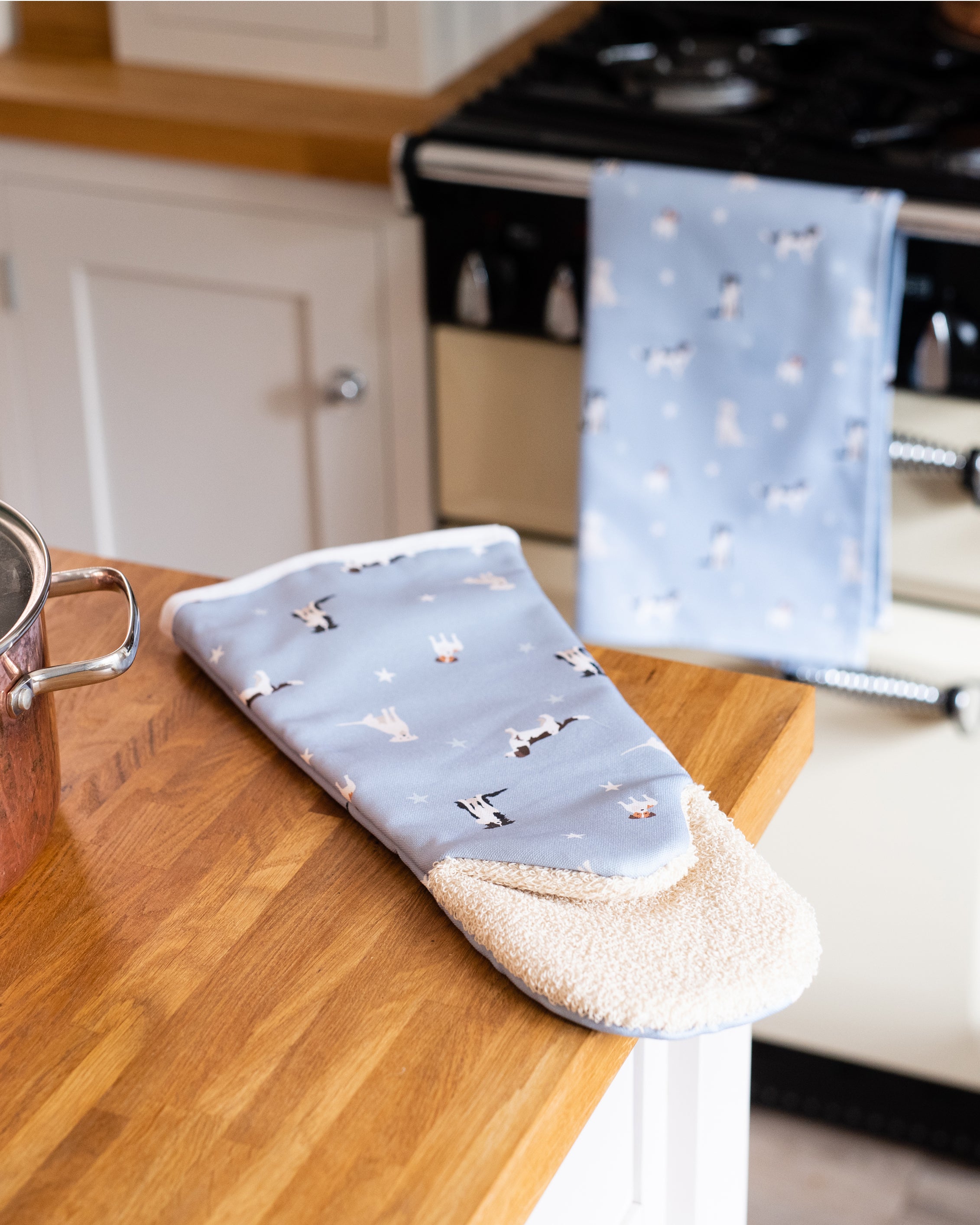Blue oven gauntlet and kitchen towel with dog pattern hanging on a stove and placed on a wooden worktop