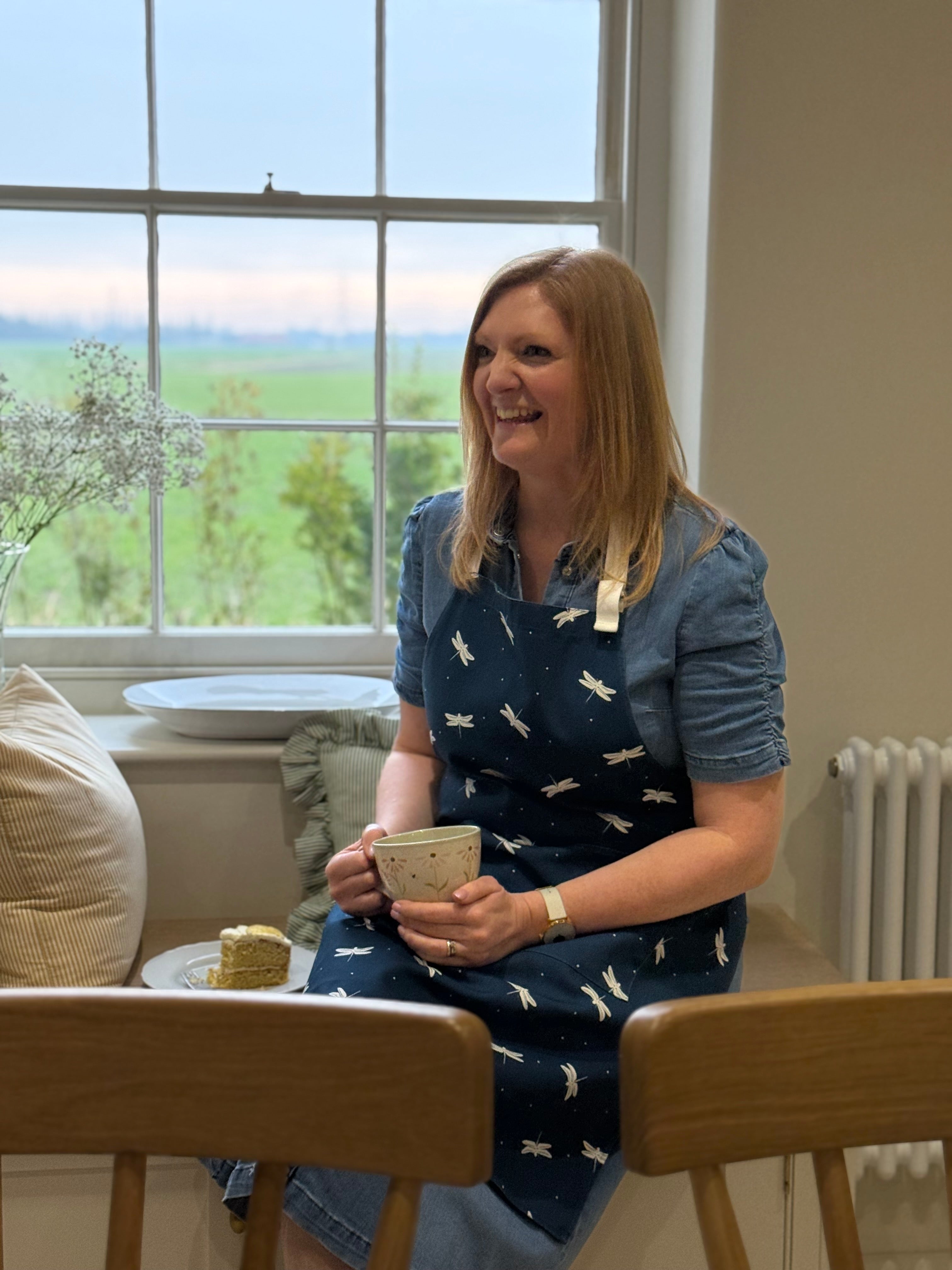 Woman wearing a navy blue dragonfly apron sitting in a window with a mug and cake