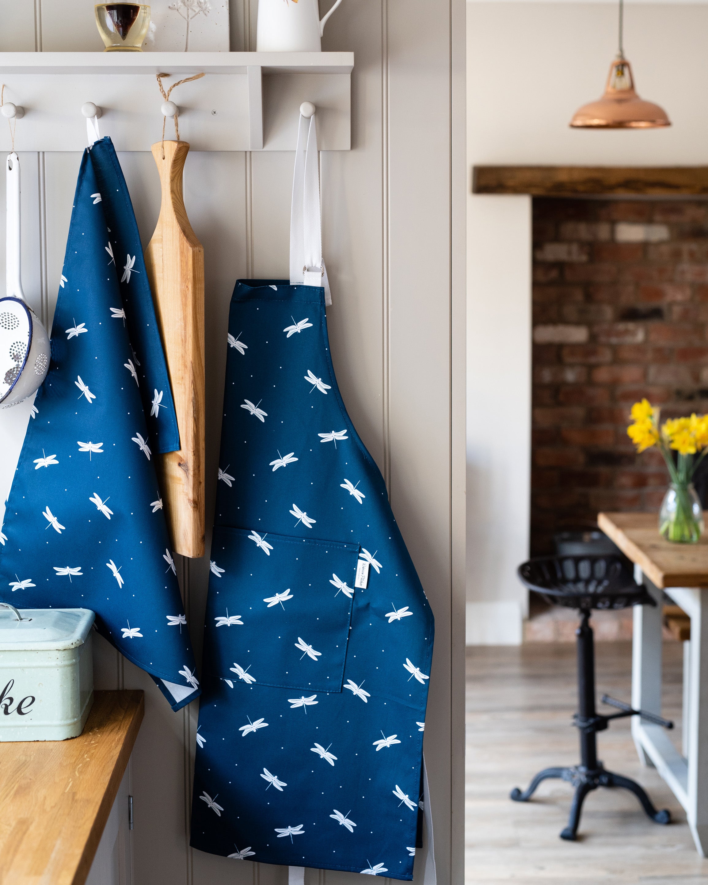 Navy blue apron and tea towel with white dragonfly pattern hanging on a wall in a kitchen.