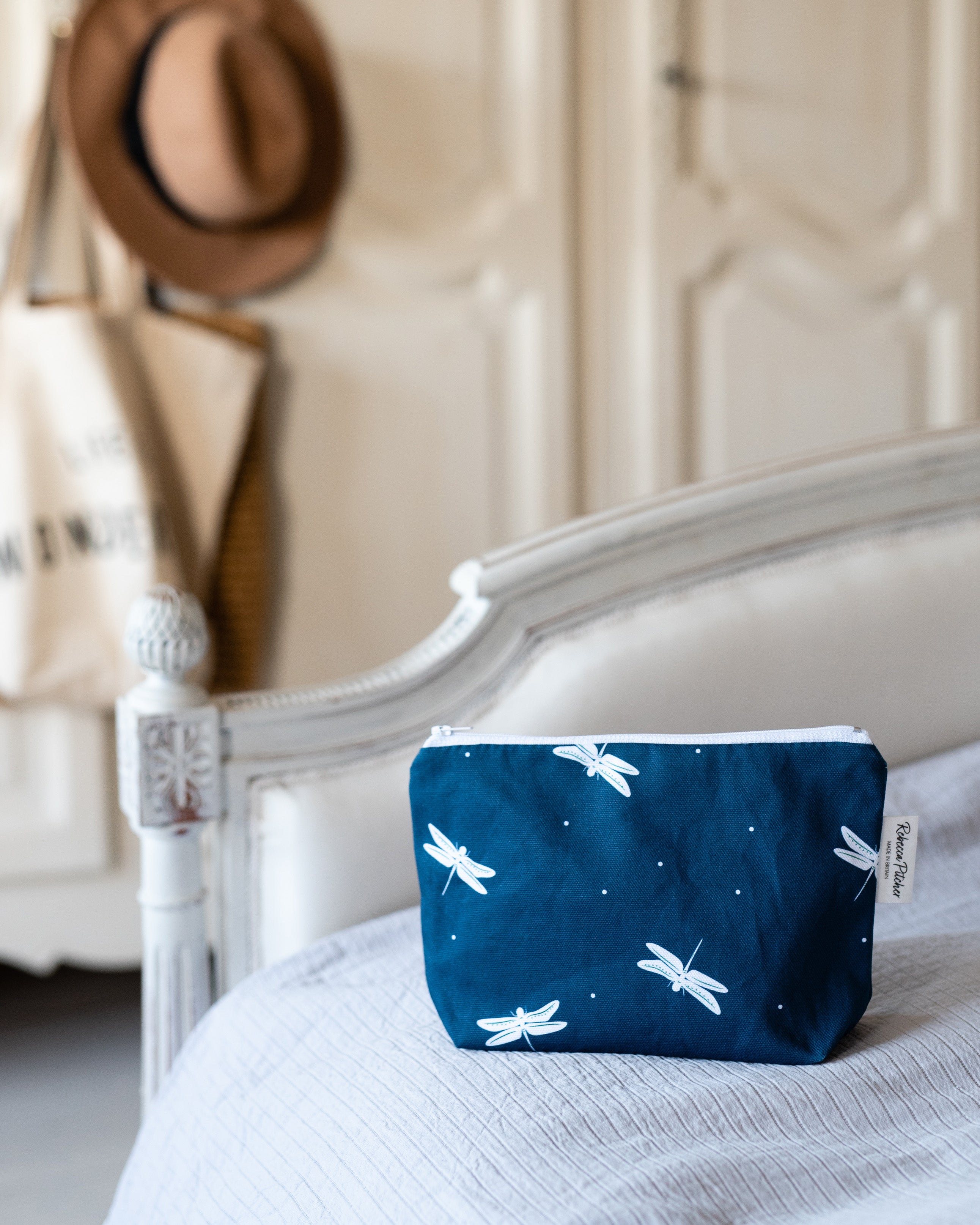 Navy blue makeup pouch with white dragonfly design on a bed with a blurred background
