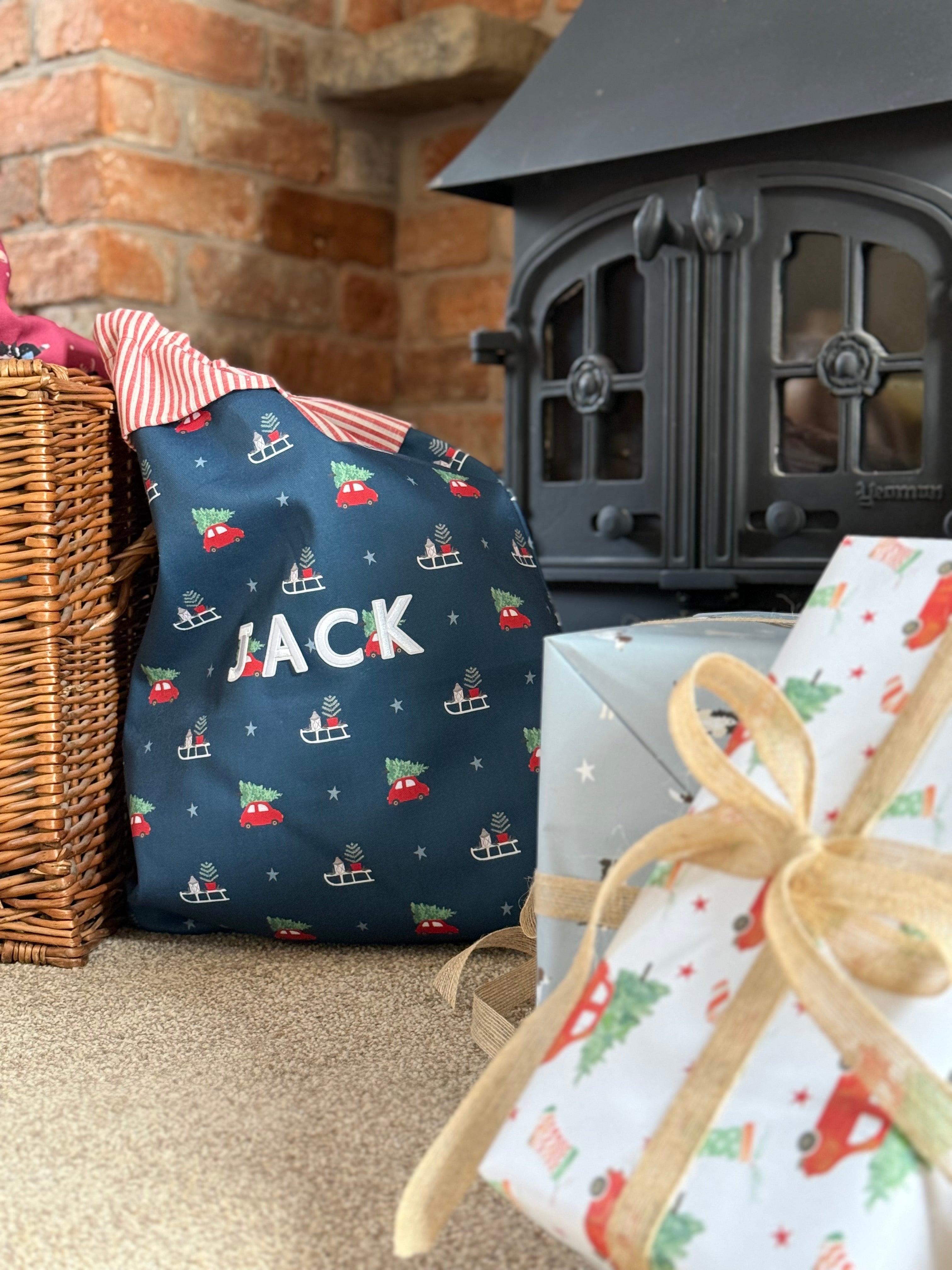 Navy personalised Santa sack with the name 'JACK' on it next to wrapped gifts in front of a fireplace.