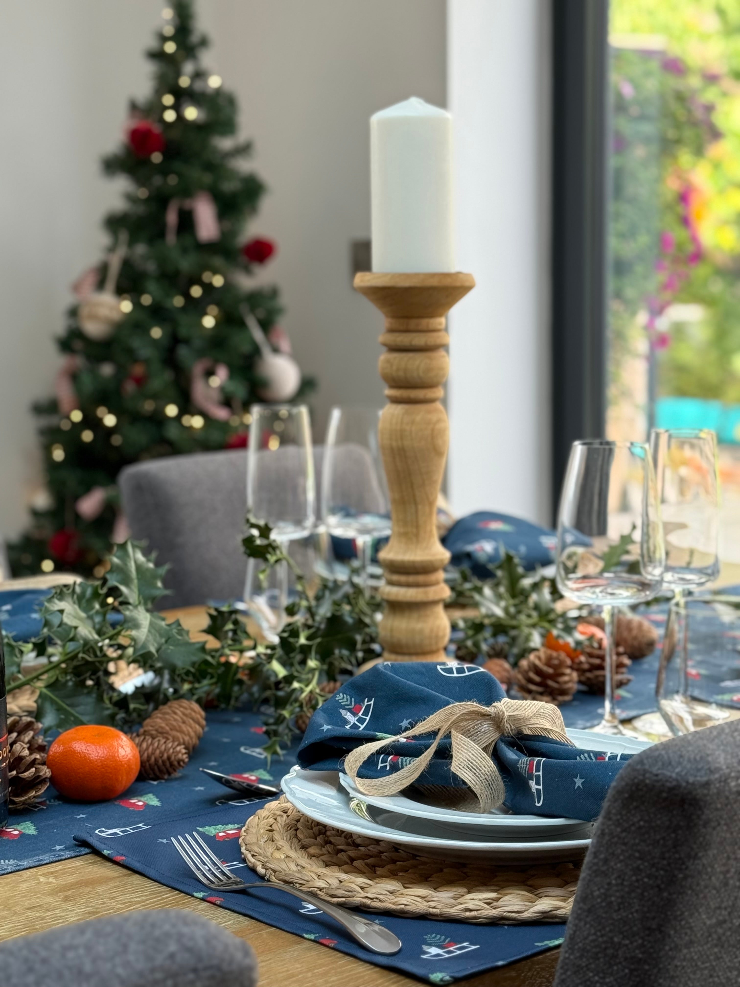 Decorated Christmas table with placemats, napkins and runner with candles, greenery, and a tree in the background.