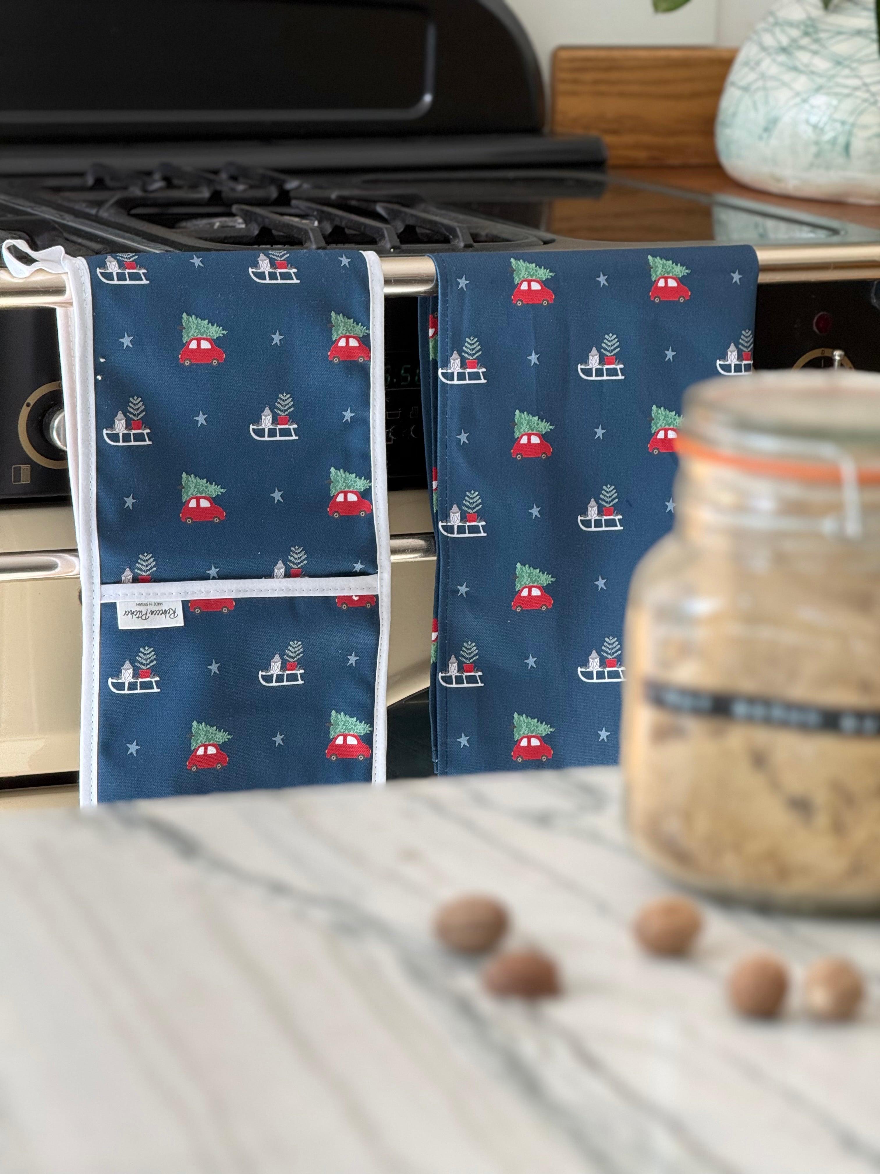 Blue ovengloves and kitchen towel with Christmas tree and sleigh pattern hanging on an oven door.