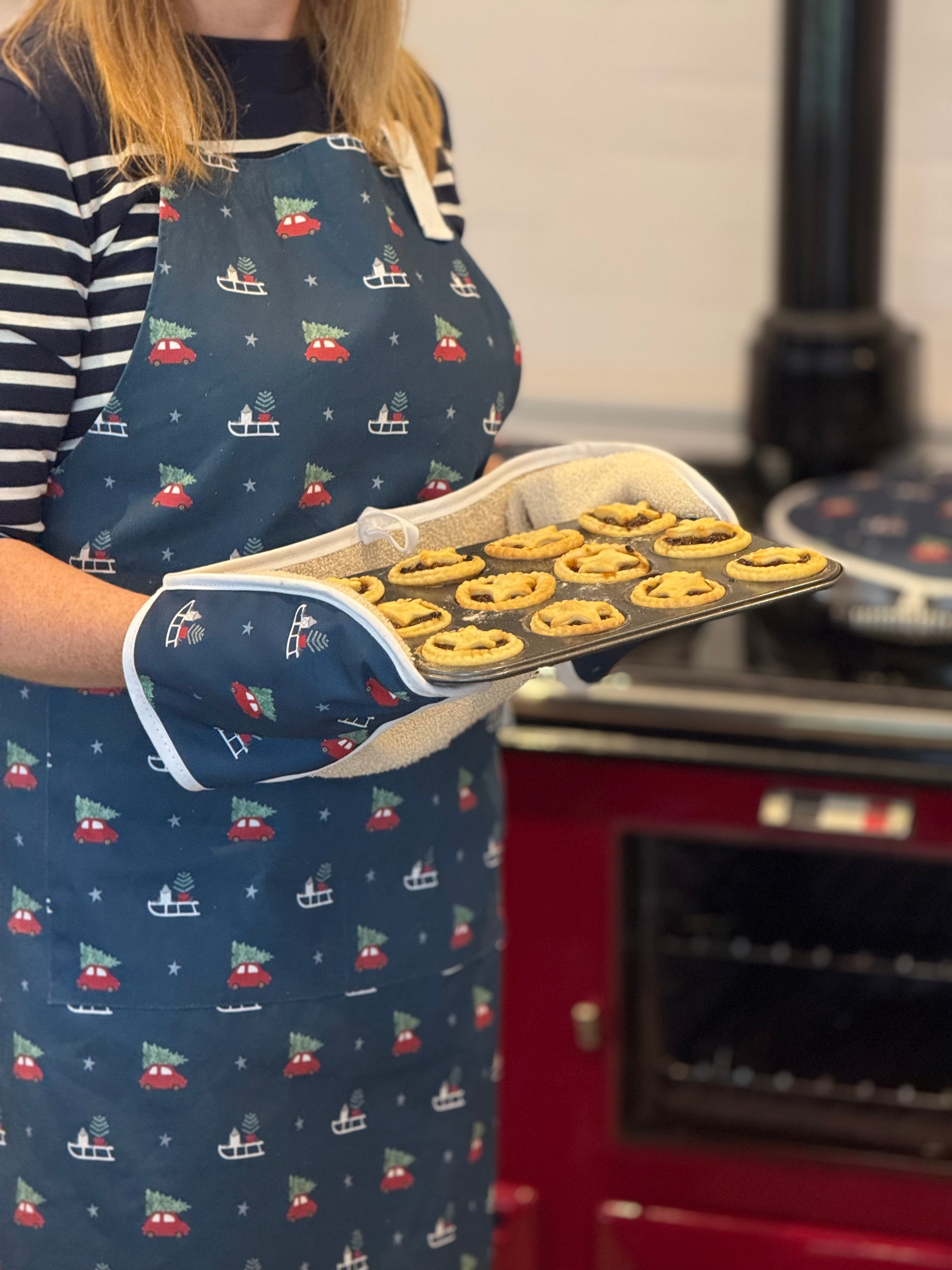 Person wearing a navy apron with a Christmas car pattern, holding a tray of mince pies in front of a red oven.