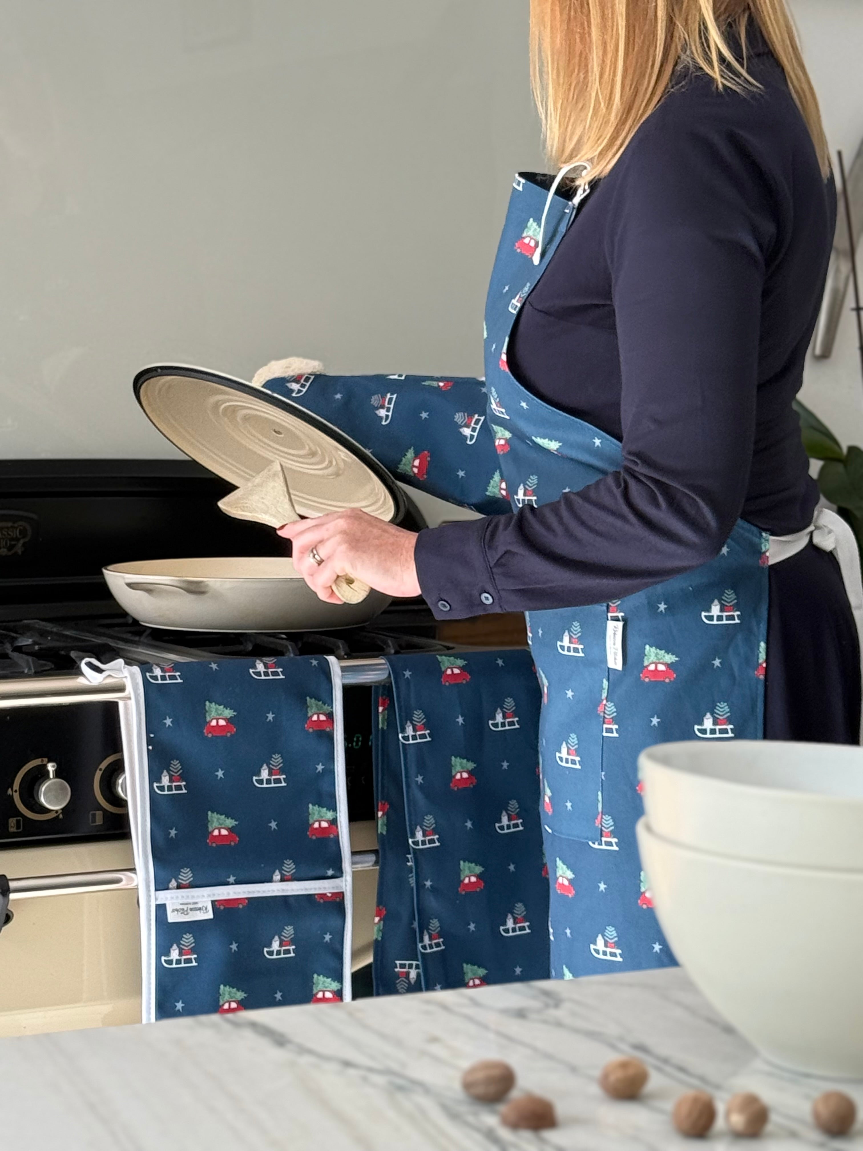 Person wearing a blue apron with Driving Home for Christmas pattern cooking at a kitchen stove.
