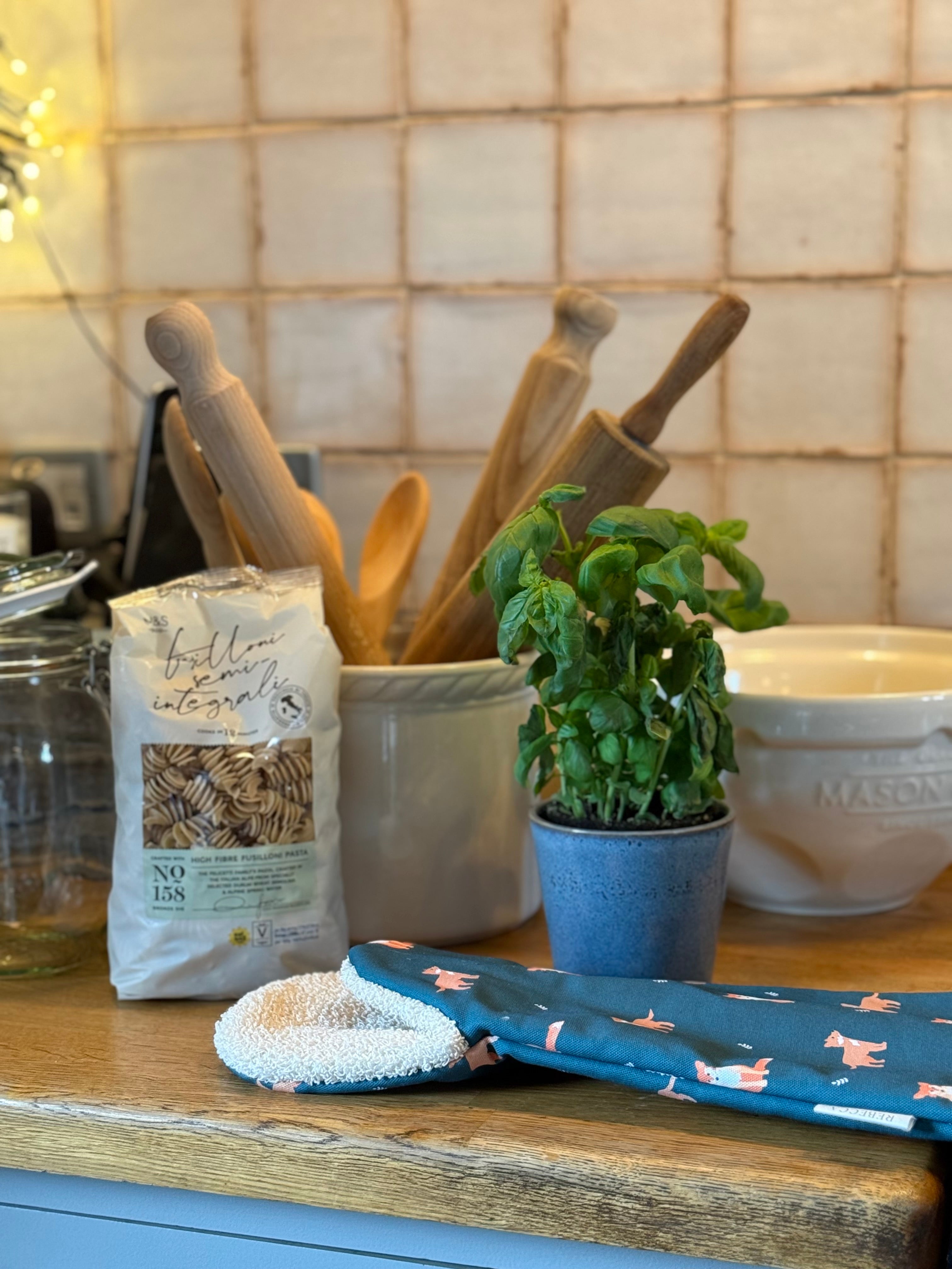 Kitchen counter with a fox design long oven gauntlet, basil plant, rolling pin, wooden spoons, and a bag of pasta