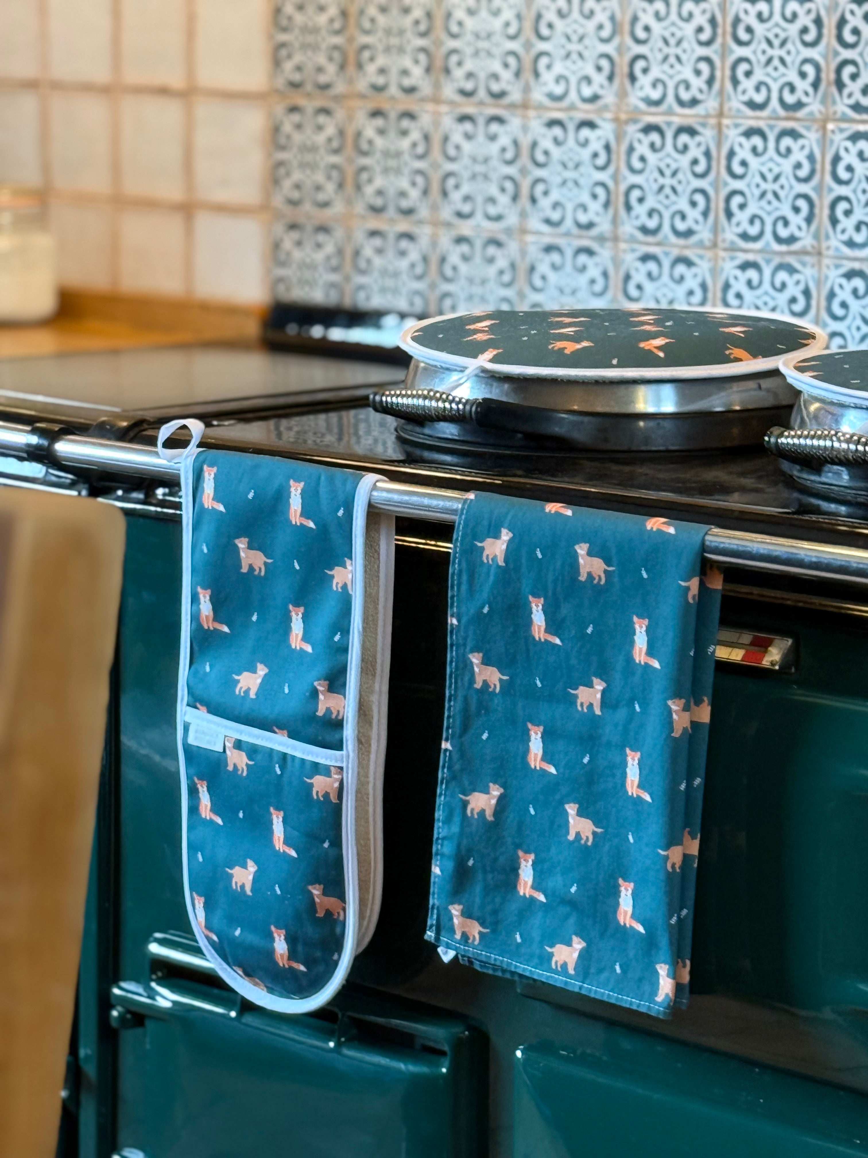 Dark green oven mitt and tea towel with fox pattern hanging on a stove in a kitchen.