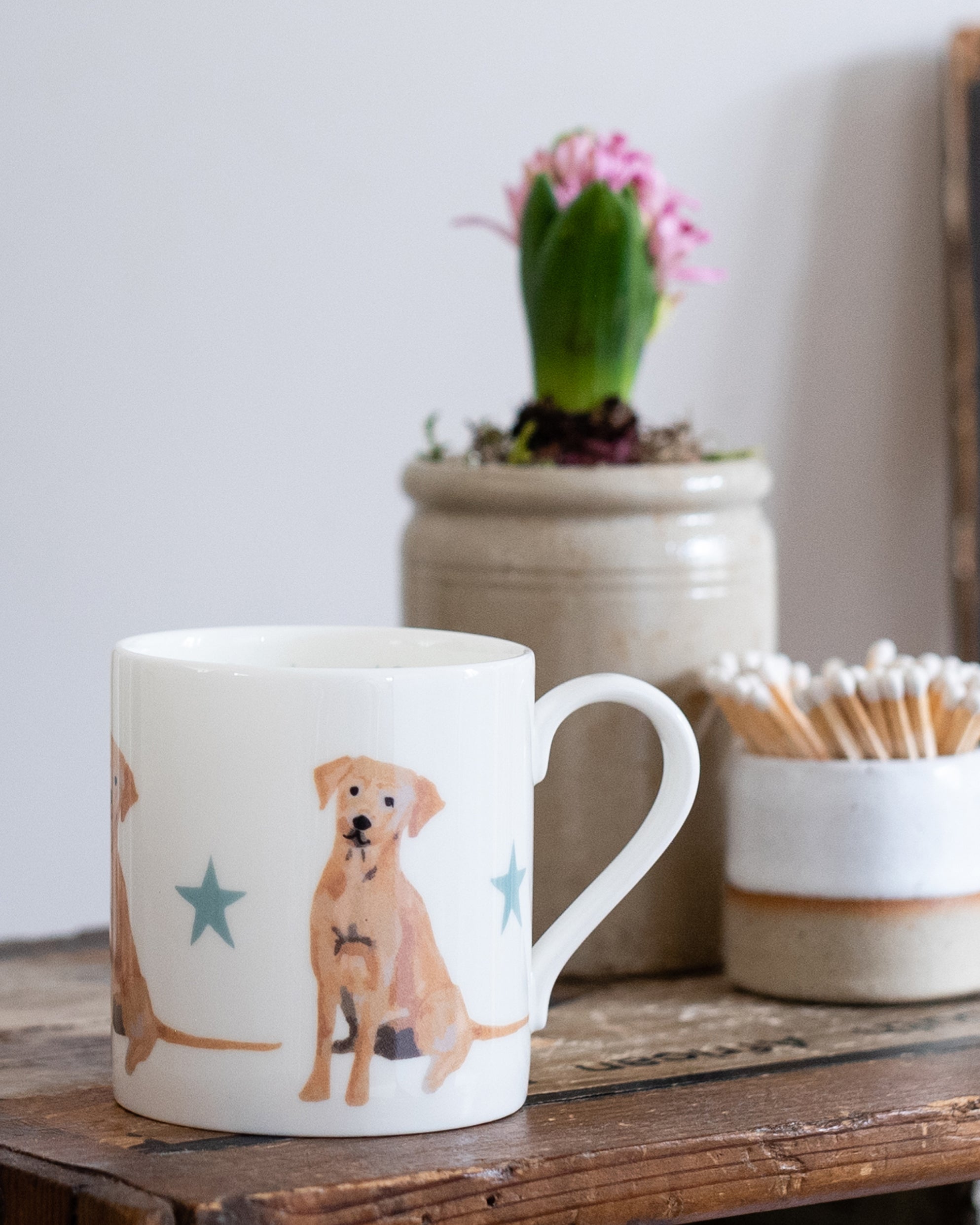 White mug with golden labrador dog illustration on a wooden surface with a plant and matches in the background