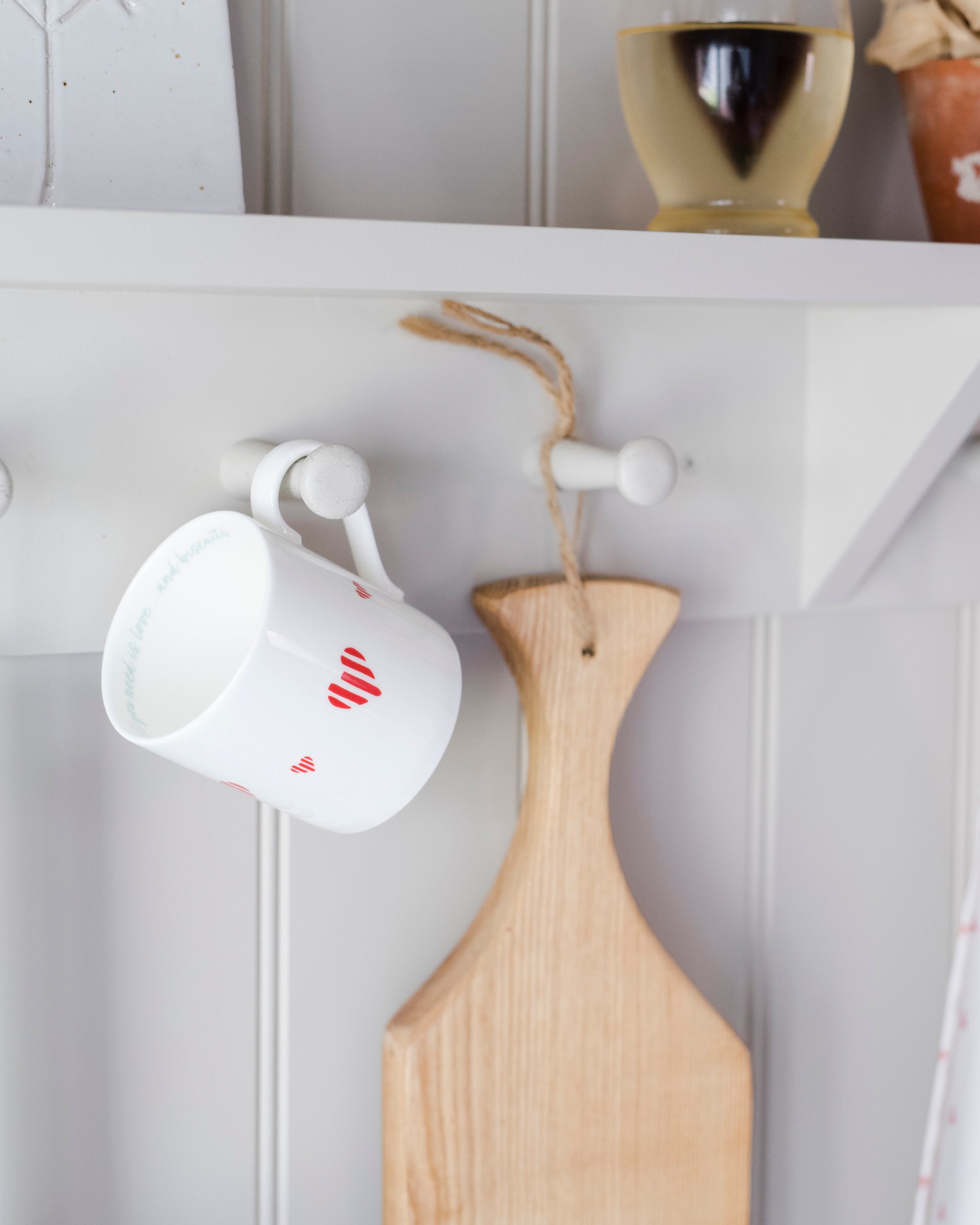 White mug with red heart design and wooden chopping board hanging on a white wall.