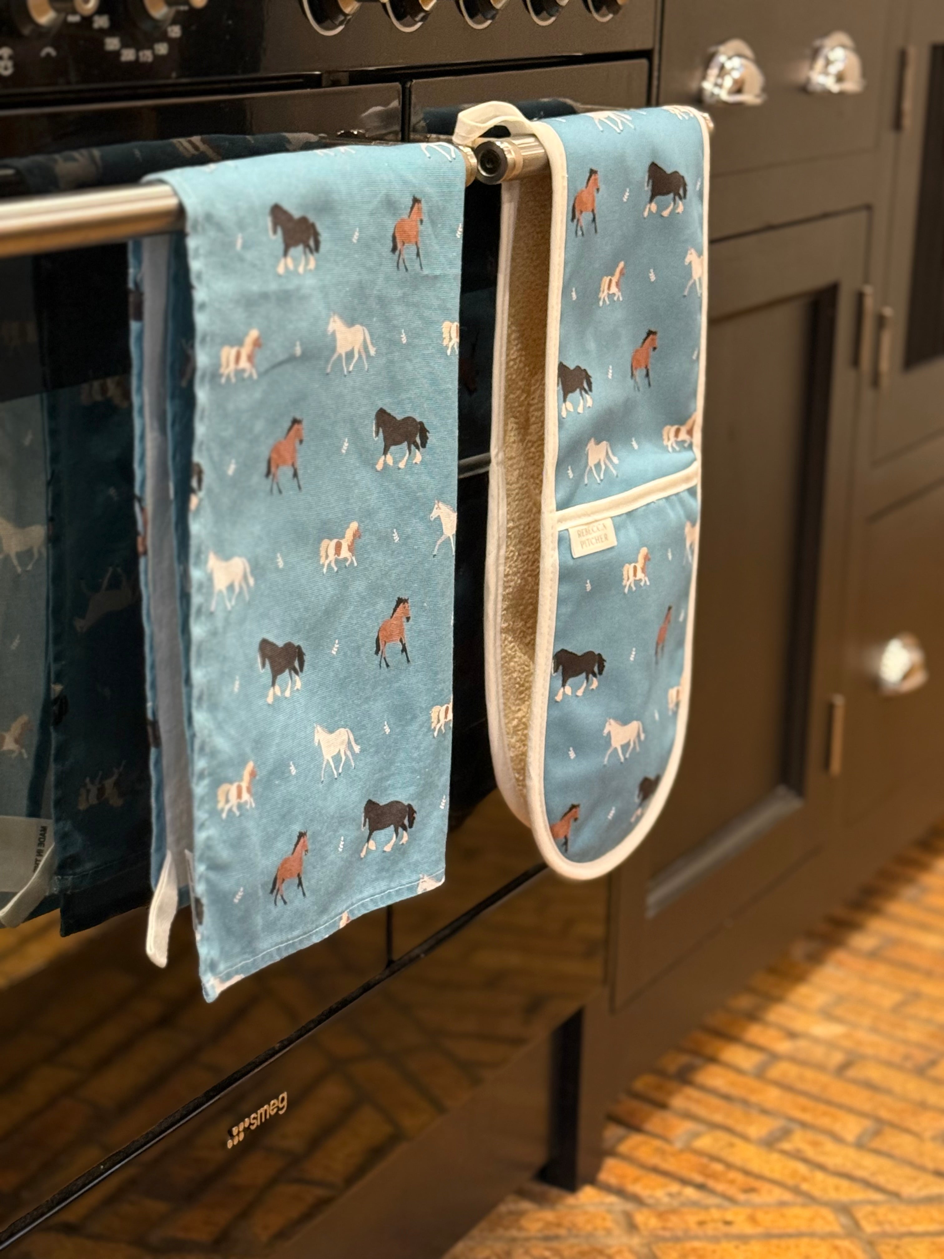 Blue tea towel and oven gloves with horse pattern hanging on a kitchen oven handle.
