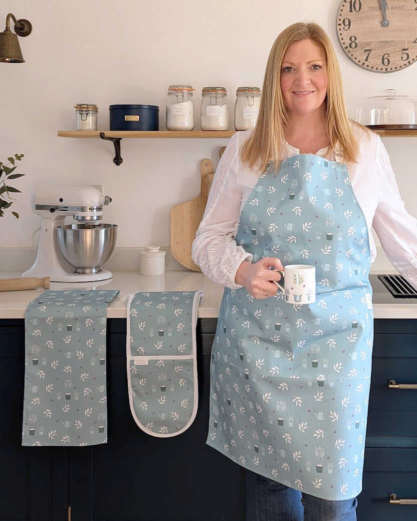 Rebecca Pitcher wearing a duckegg blue cottage garden design apron with kitchen items in a home kitchen setting