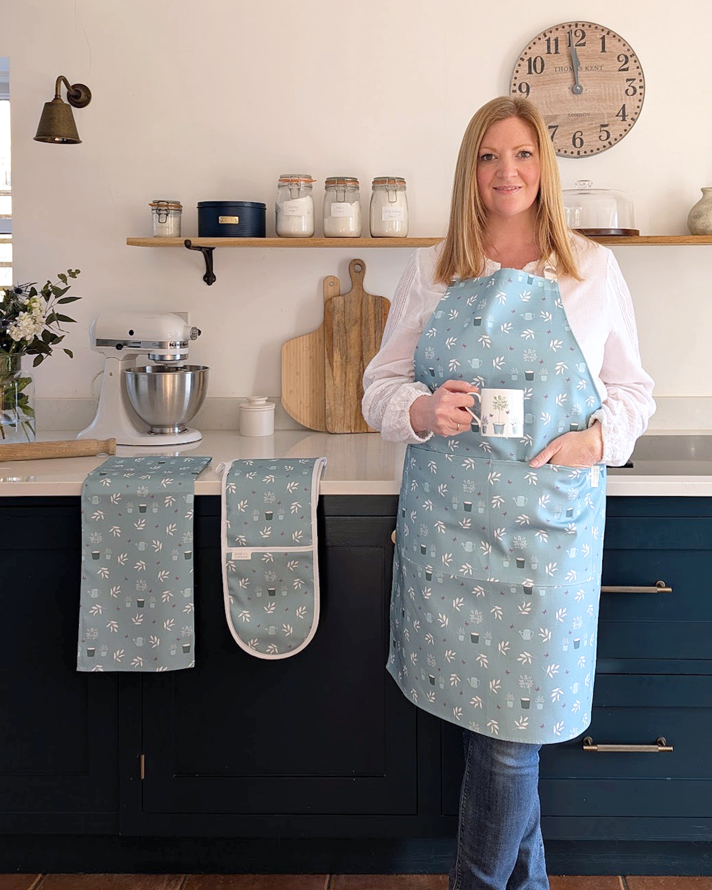 Rebecca Pitcher in a kitchen wearing a light blue apron with floral patterns, holding a mug.