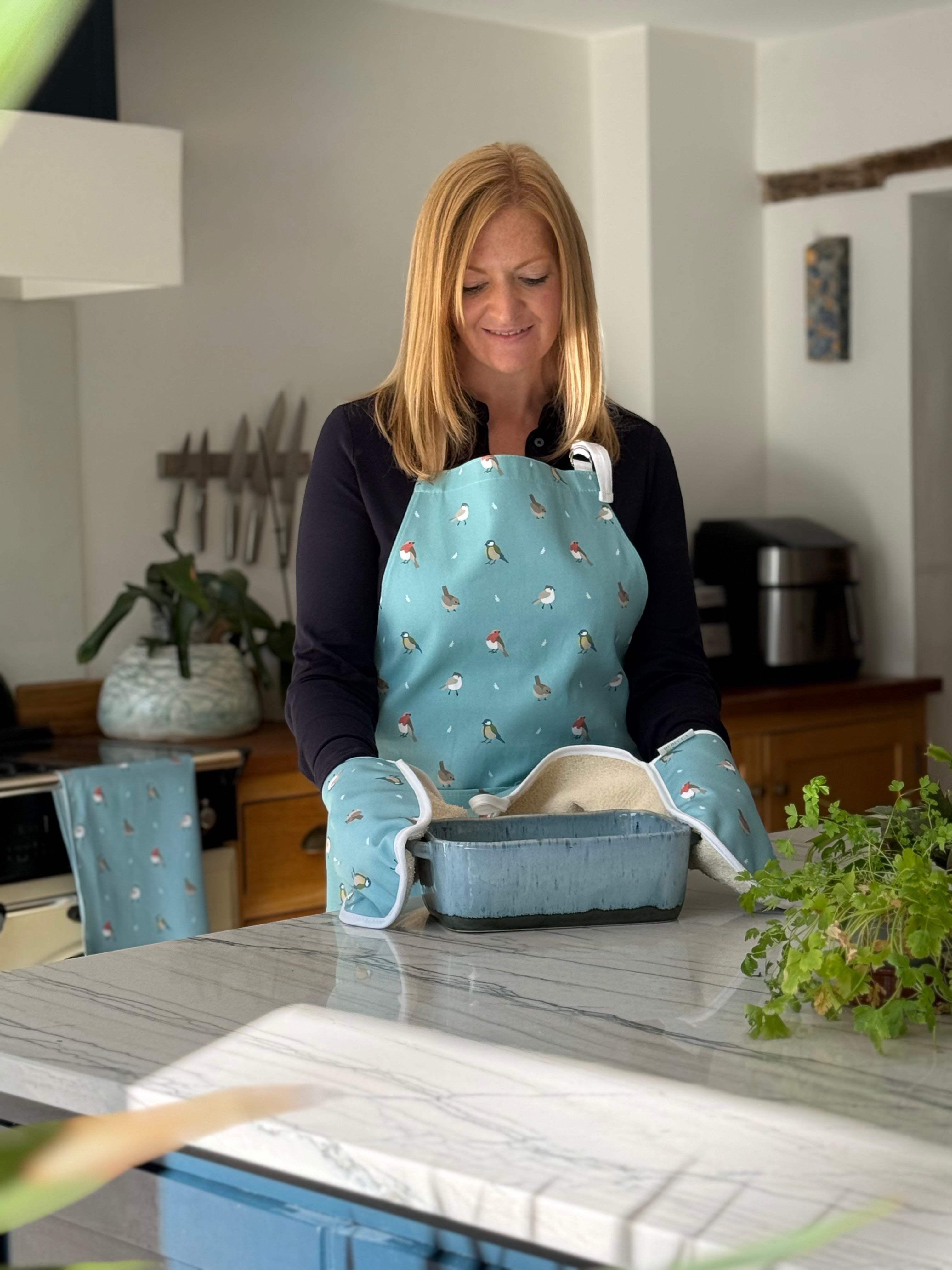 Rebecca Pitcher in a kitchen wearing an apron and oven mitts, holding a baking dish.