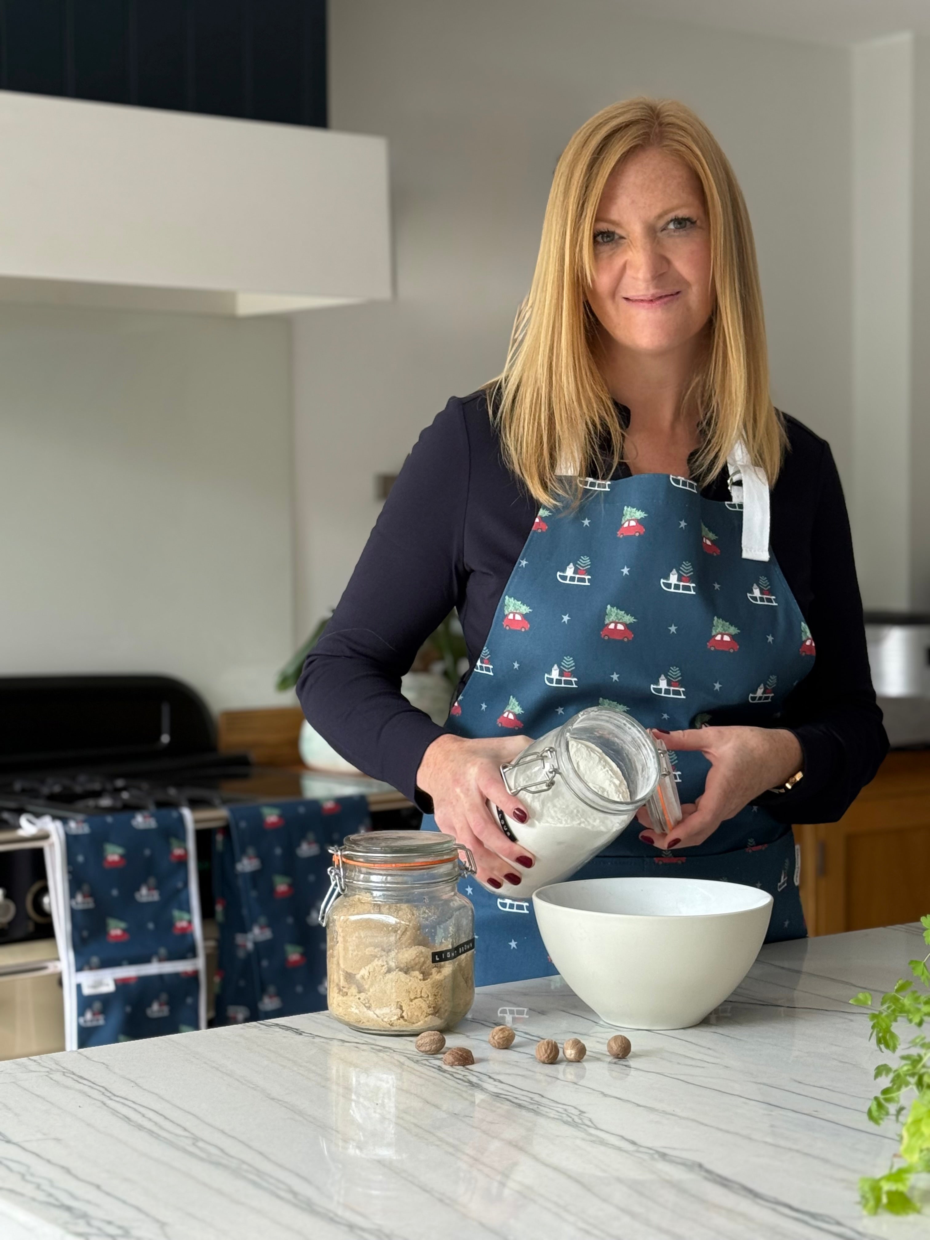 Rebecca Pitcher in a Chistmas apron in a kitchen pouring ingredients from a jar into a bowl