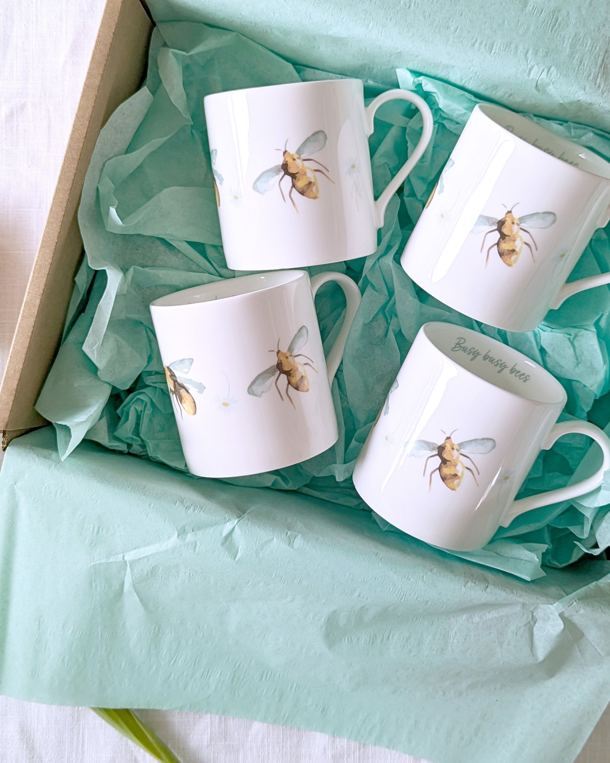 Four white mugs with bee designs in a box lined with green tissue paper.