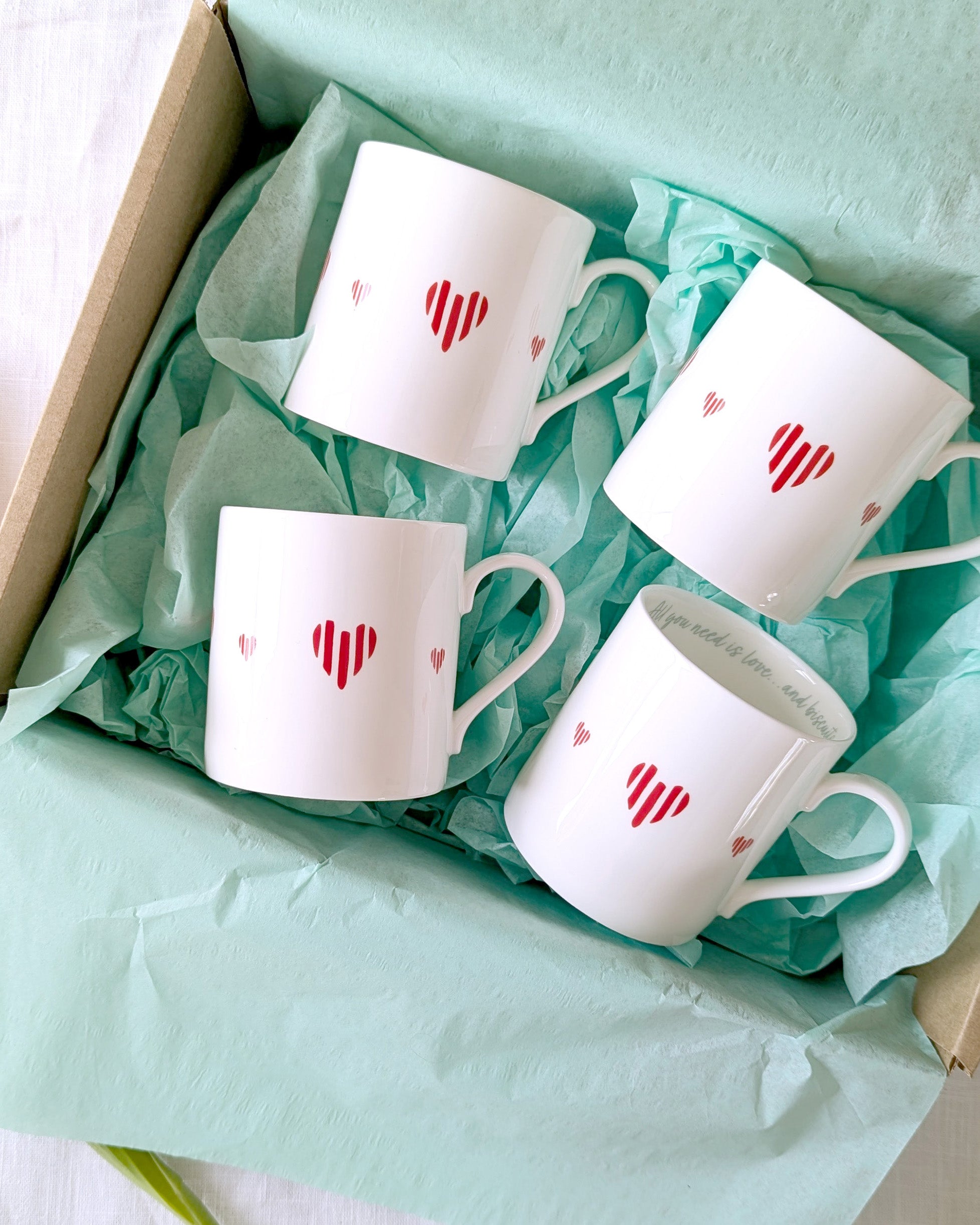 Four white mugs with red heart designs in a box lined with green tissue paper.