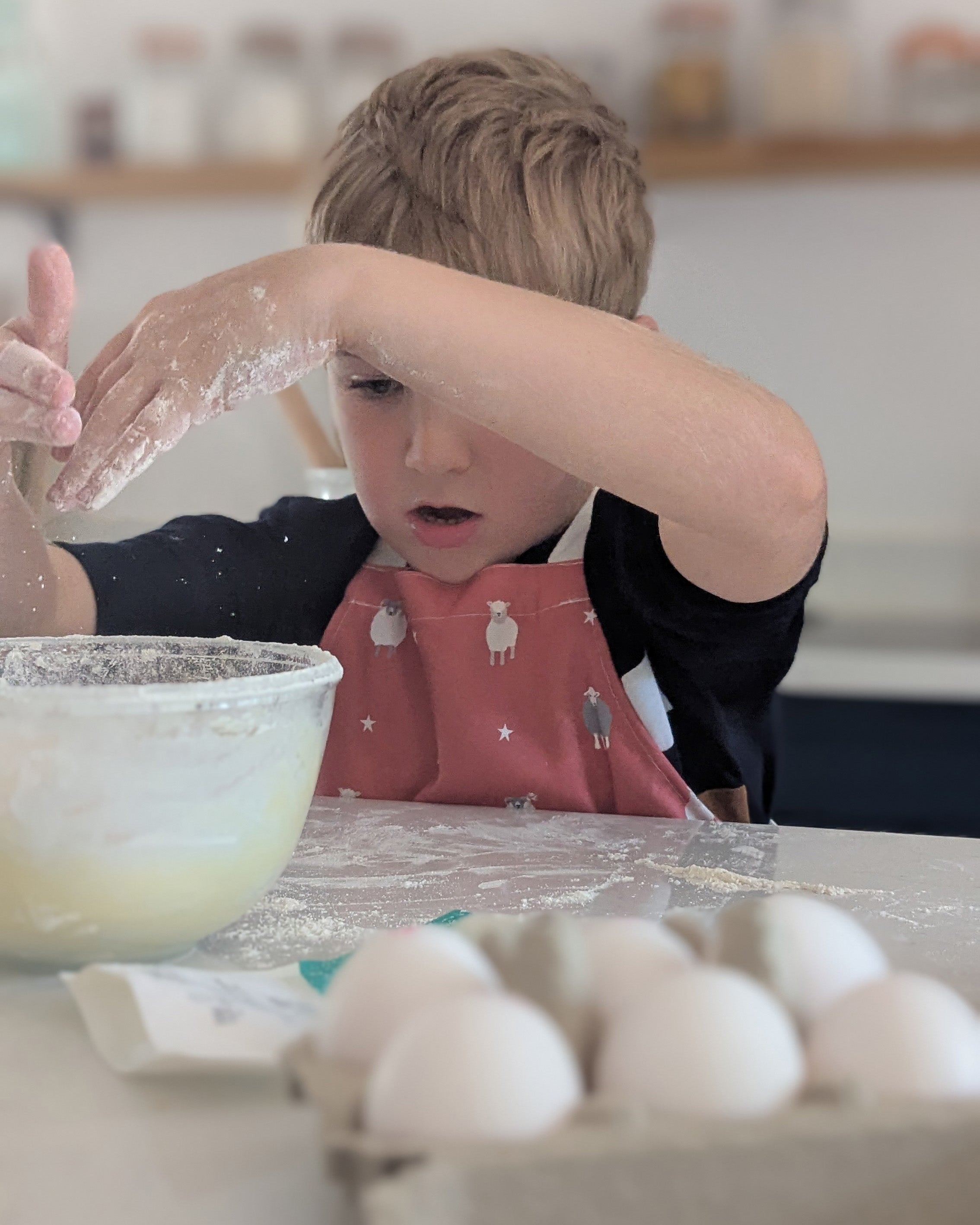 Child wearing a red sheep design apron in a kitchen with flour on hands and apron, near eggs and a bowl.