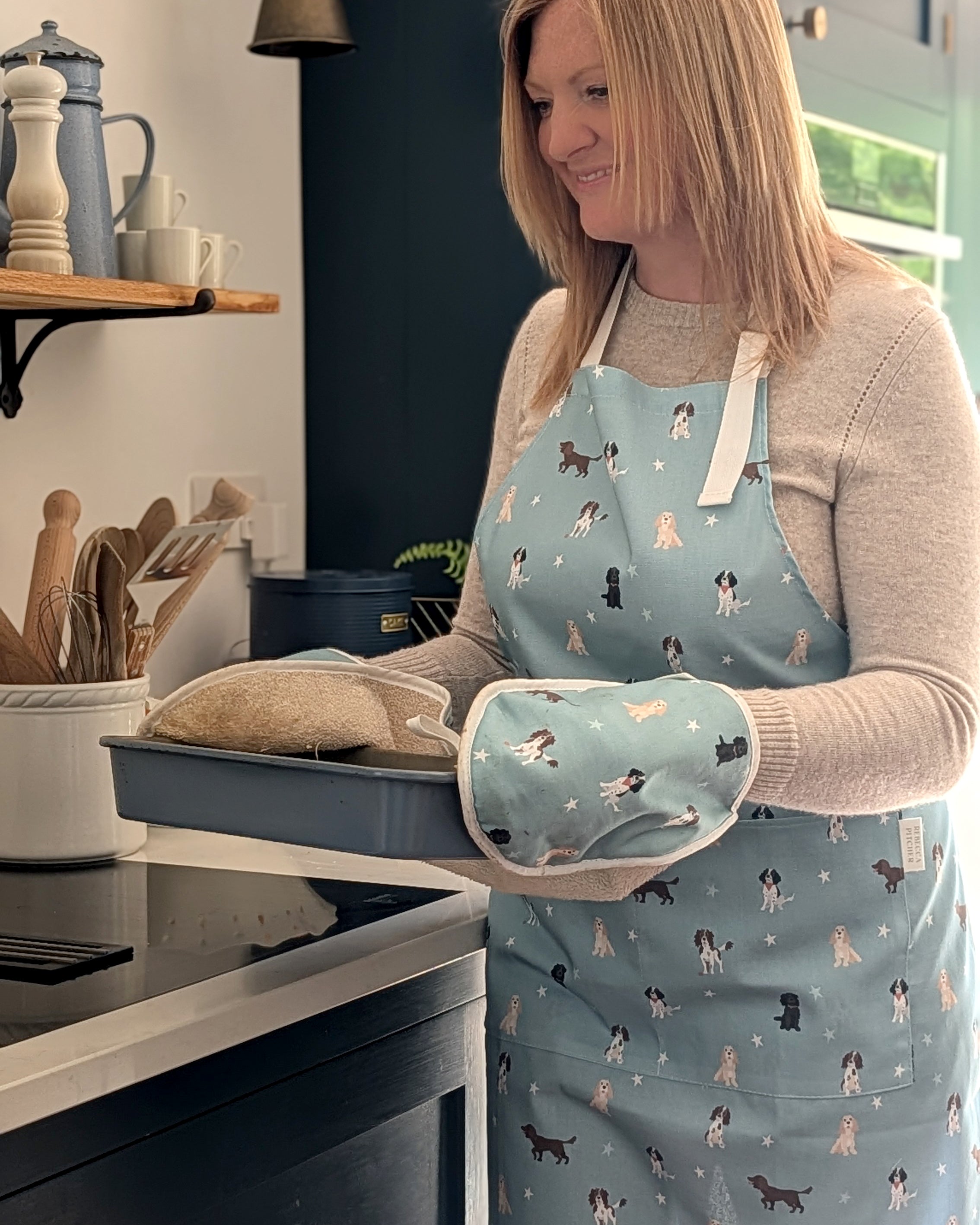 Woman in a kitchen wearing a blue apron with dog patterns, holding an oven mitt and pan.