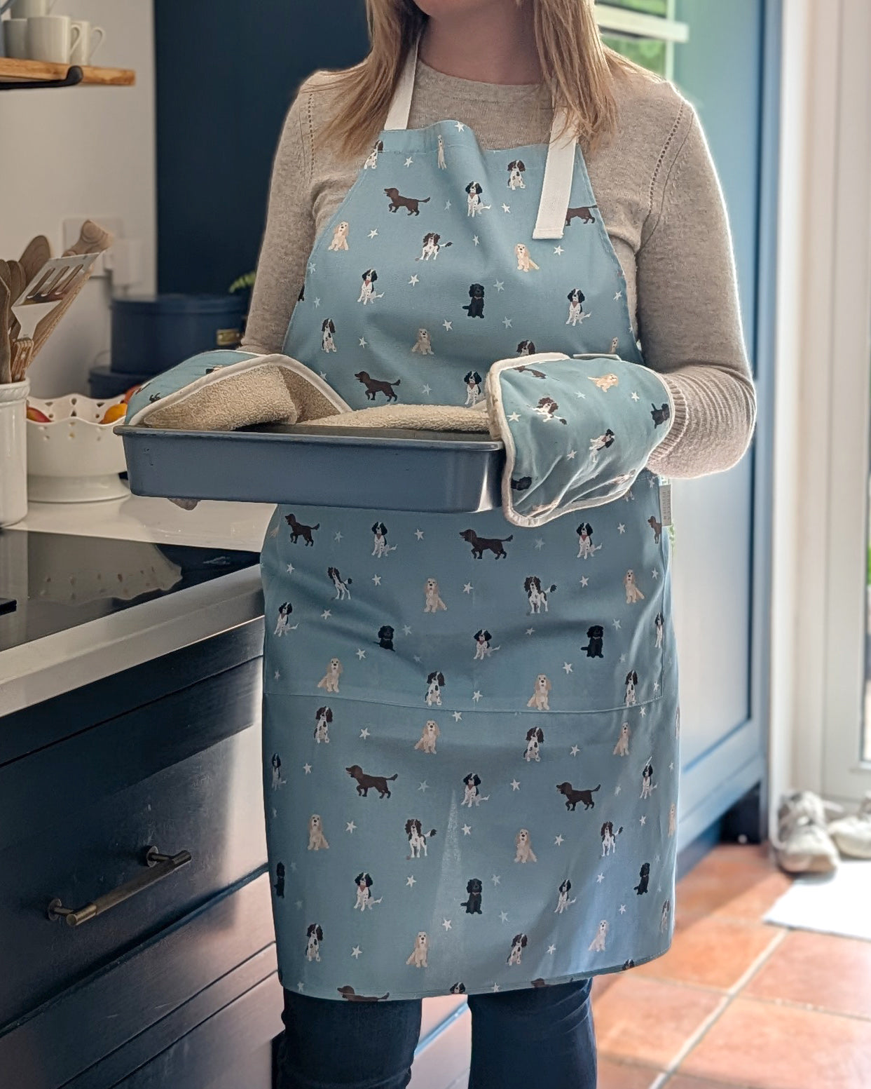 Person wearing a blue apron with dog pattern holding a baking tray in a kitchen.