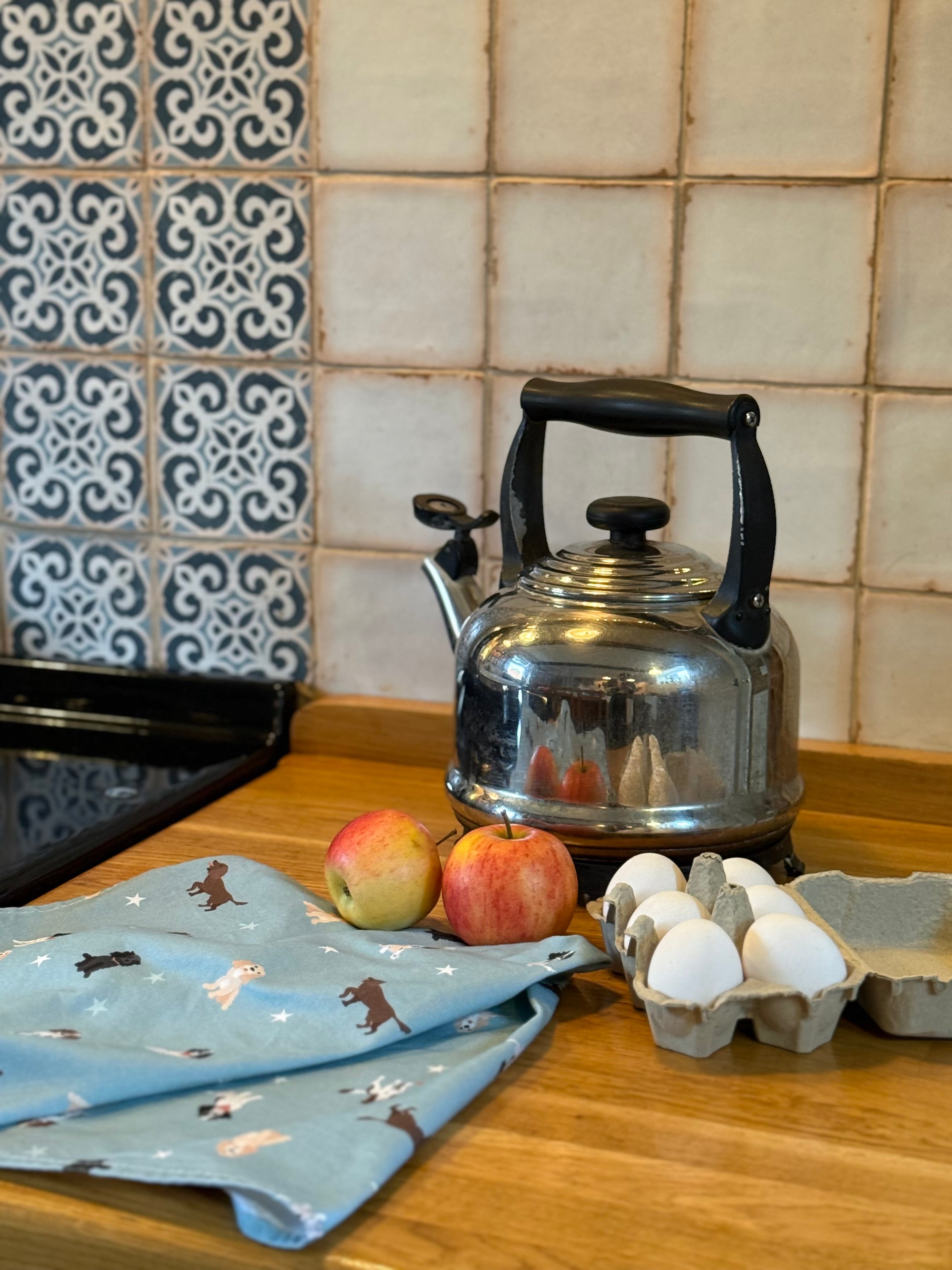 Blue spaniel dog patterned tea towel in front of a traditional kettle on a wooden counter against a tiled wall.